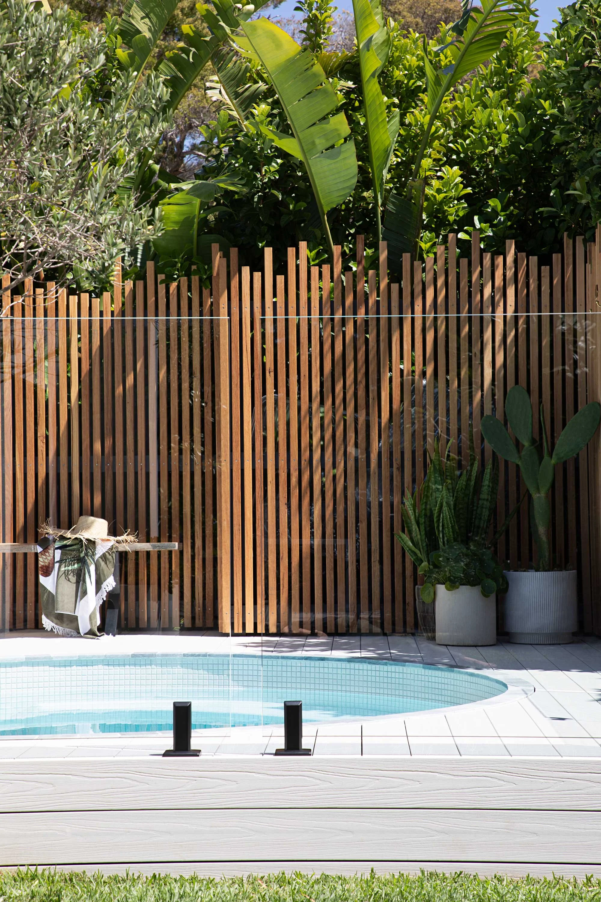 Backyard with a swimming pool, a wooden fence, potted plants, a bench seat and beach towel, and lush greenery.