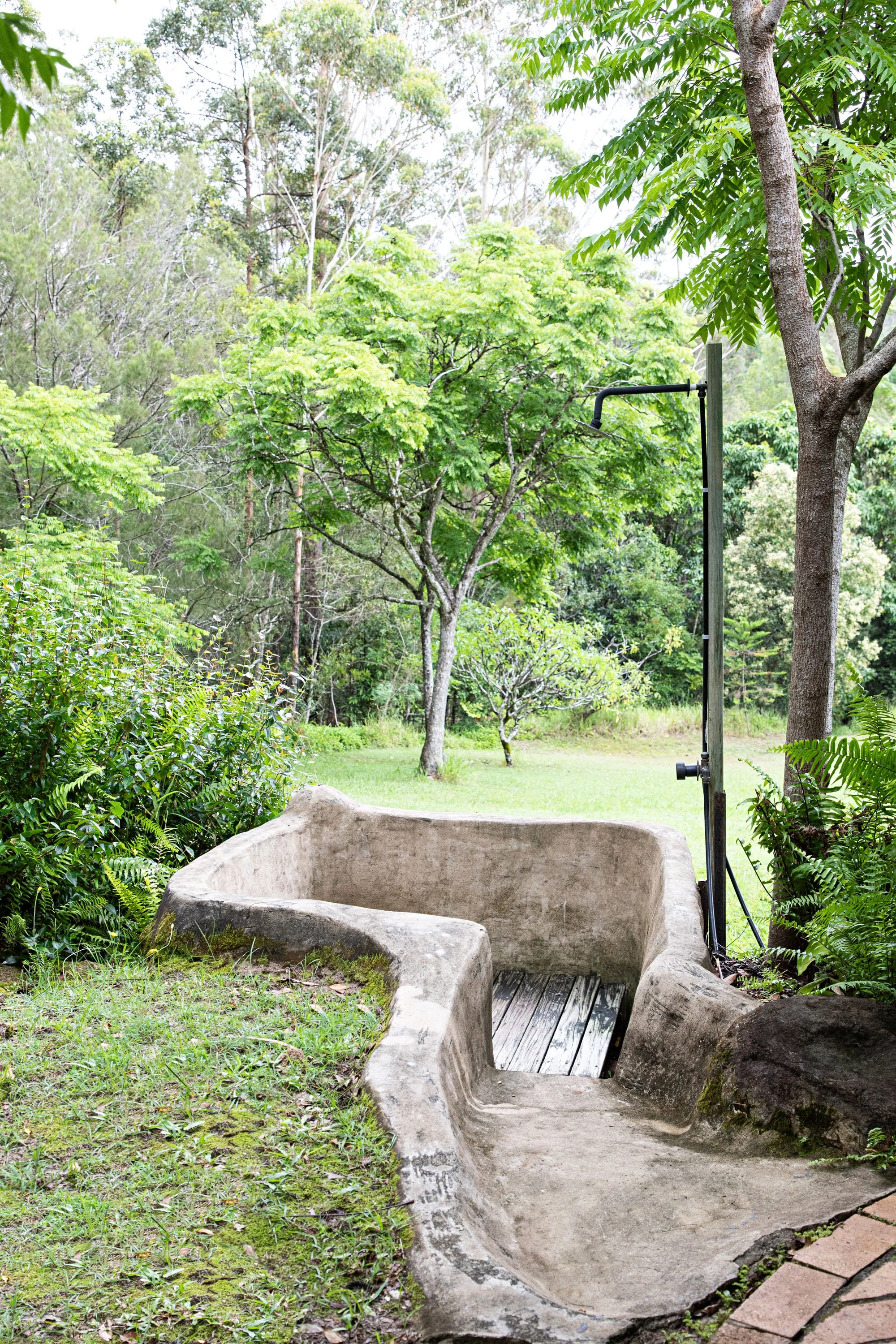 A stone water fountain with a built-in spout, surrounded by greenery and trees in a garden.