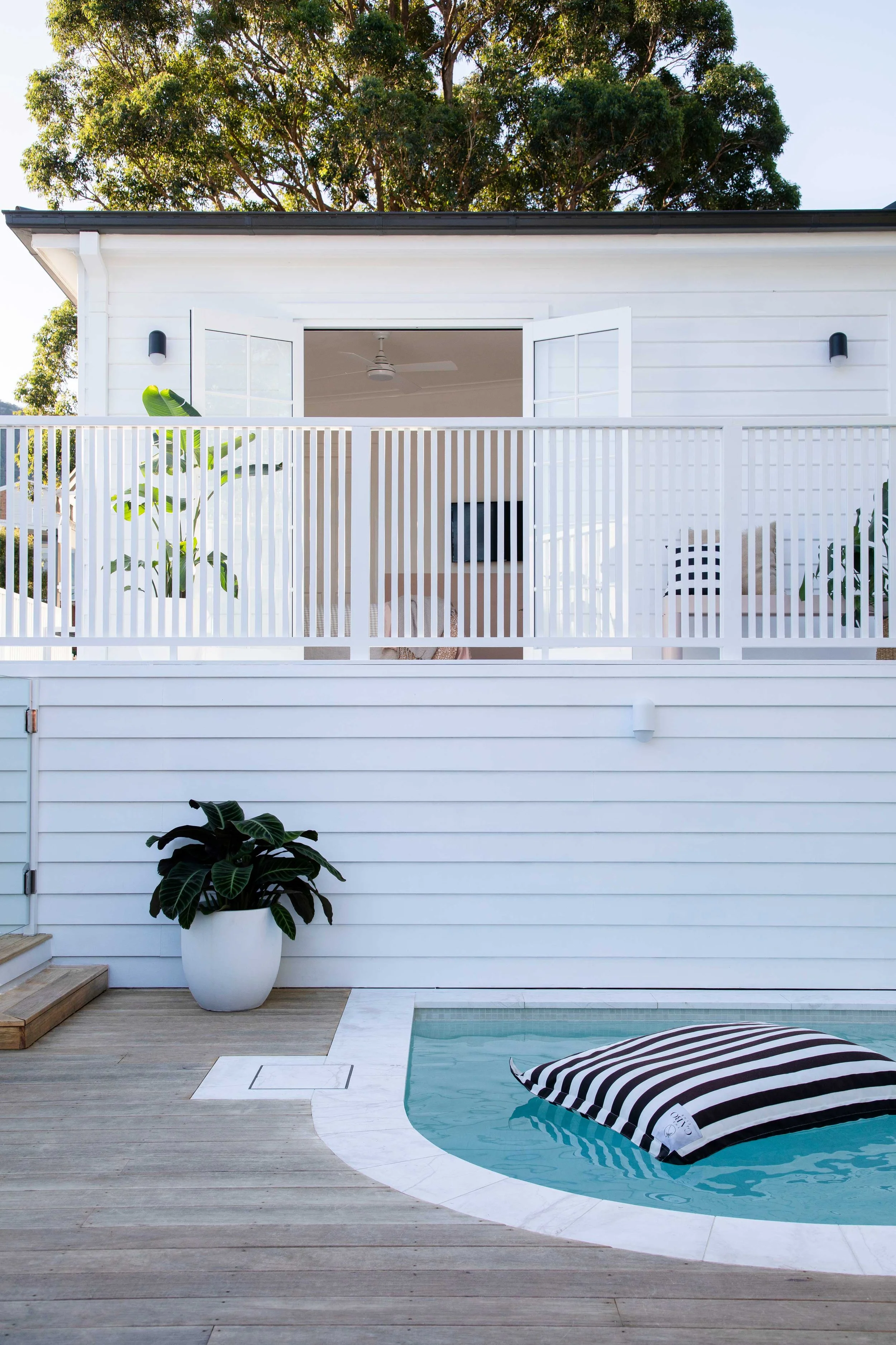 Residential backyard with small swimming pool, striped inflatable float, wooden deck, white fence, tropical potted plant, and white house exterior with open balcony and ceiling fan.