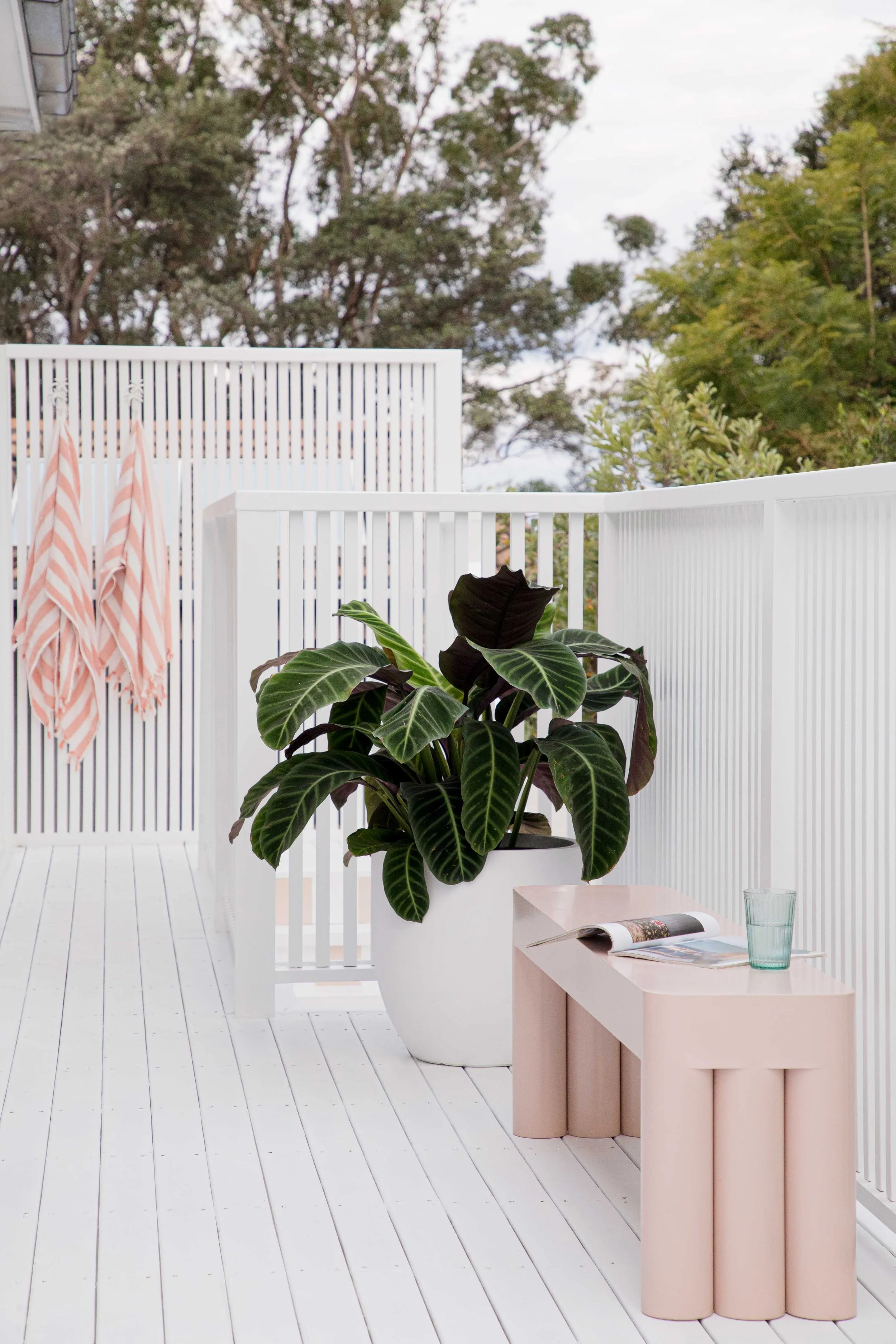 A white outdoor deck with a white railing and white floorboards, featuring a large potted plant with broad dark green leaves on a white stand, a pink decorative table with a scalloped design, a glass of water, and an open magazine, with trees and a c