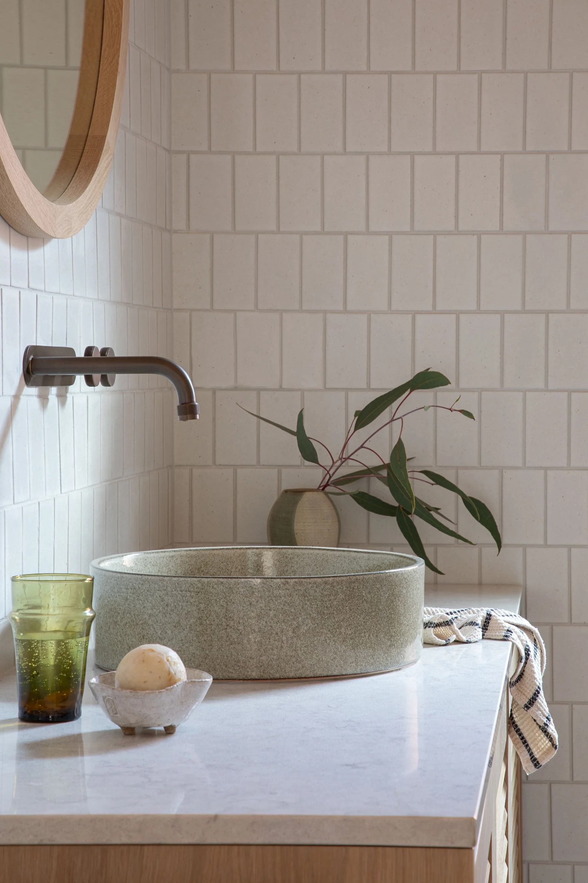 A minimalist bathroom vanity with a rounded stone sink, a clay-colored countertop, and a small potted plant with green leaves. A striped towel hangs on the side, and there is a glass of water and a soap in a dish nearby. The background features white
