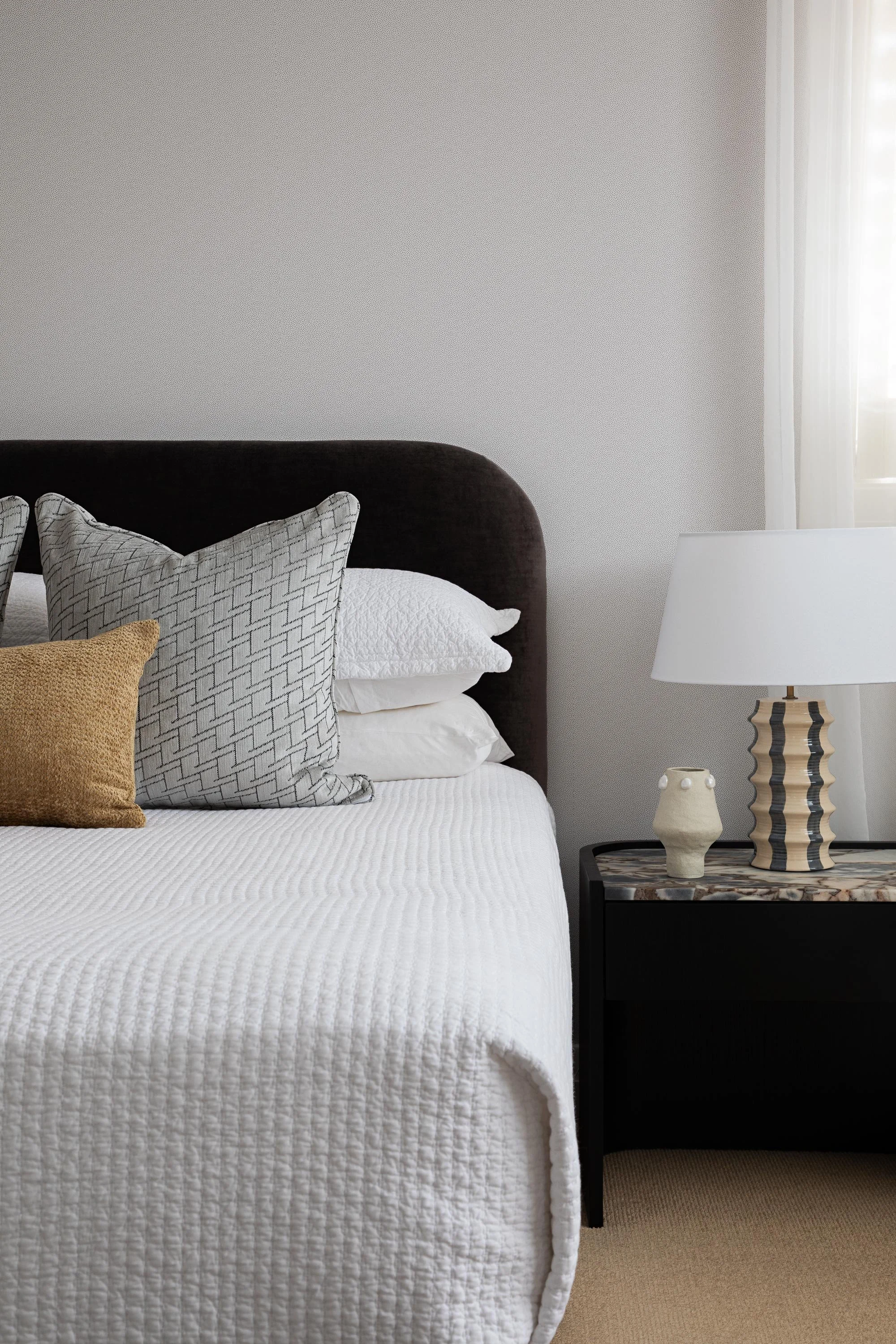 A neatly made bed with a white quilt and multiple decorative pillows in front of a plain wall. To the right is a black nightstand with ceramic vases and a striped table lamp, with sunlight coming through sheer curtains.