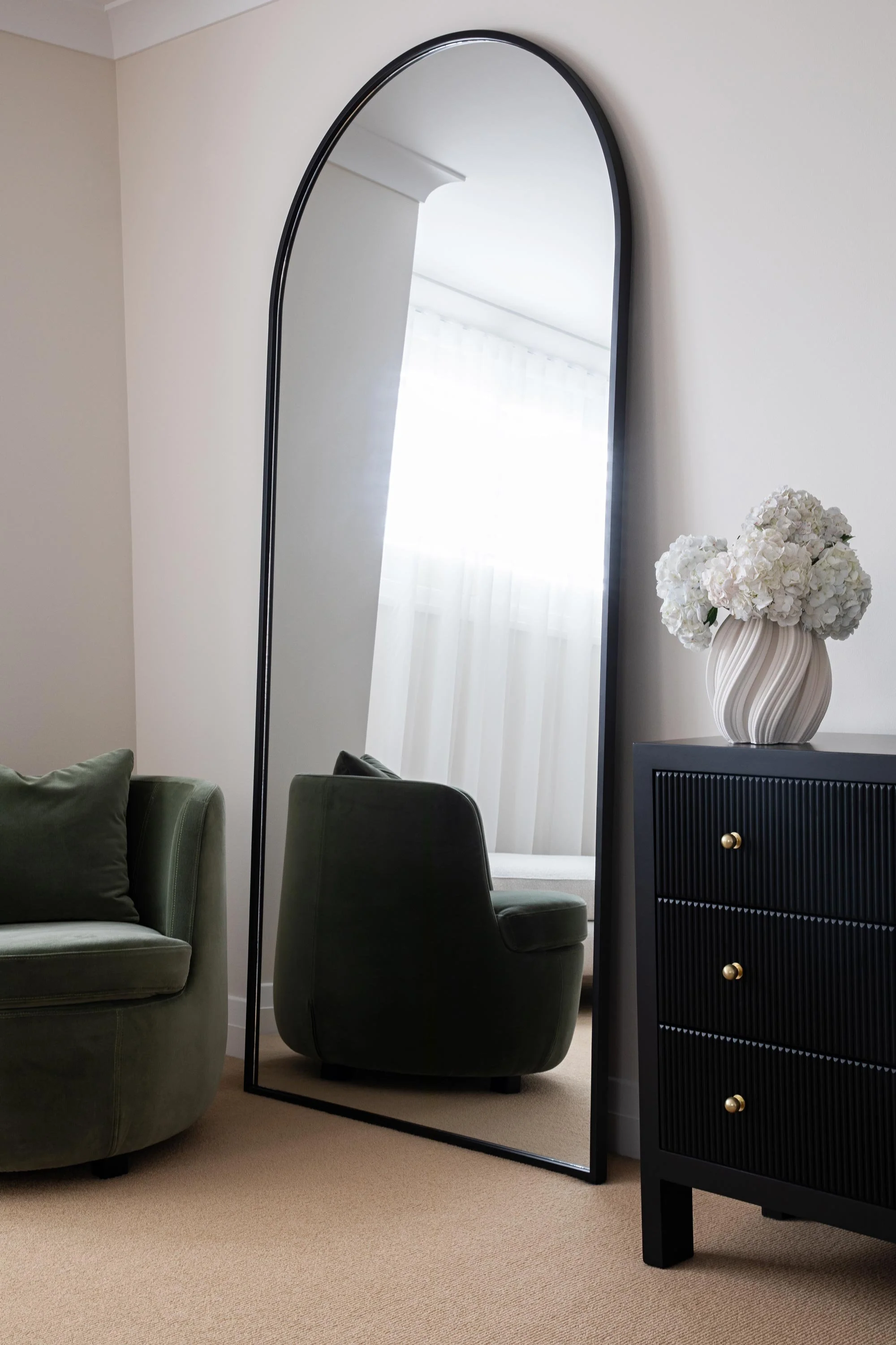 A room with beige carpet, a black dresser with three drawers and gold knobs, a white vase with white hydrangeas, a tall black-framed mirror, and green armchairs with cushions, a window with sheer curtains, and a white ceiling.