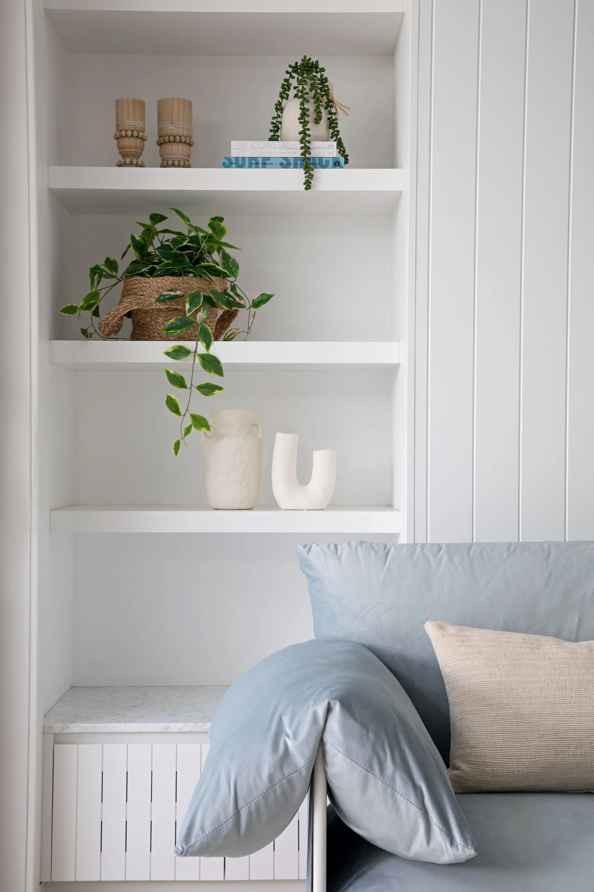 Interior of a built-in white bookshelf with decorative items, including potted plants, candles, and books, against paneling wall, with a bed and pillows in front.