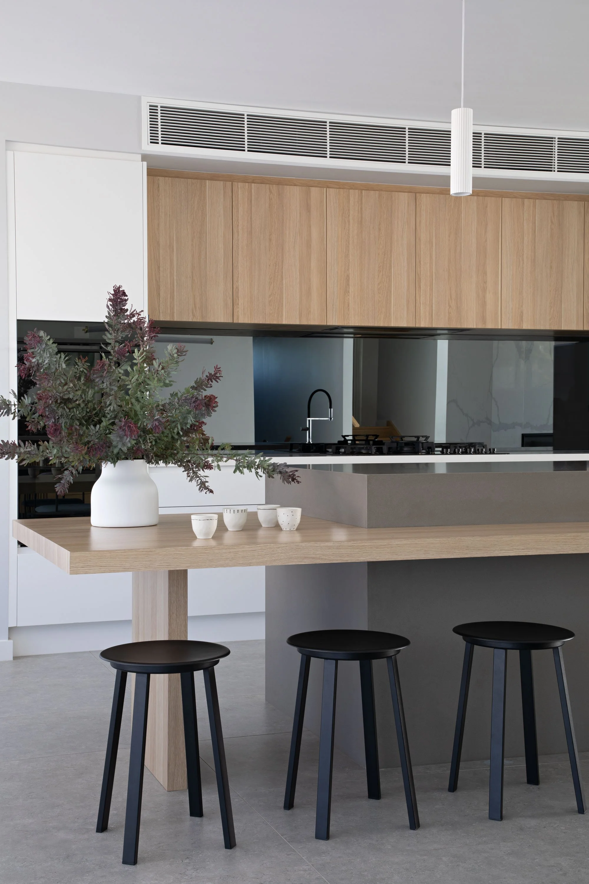 Modern kitchen with a wooden table, black bar stools, a white vase with purple and green foliage, and a sleek black coffee machine, all under a minimalist white pendant lamp.