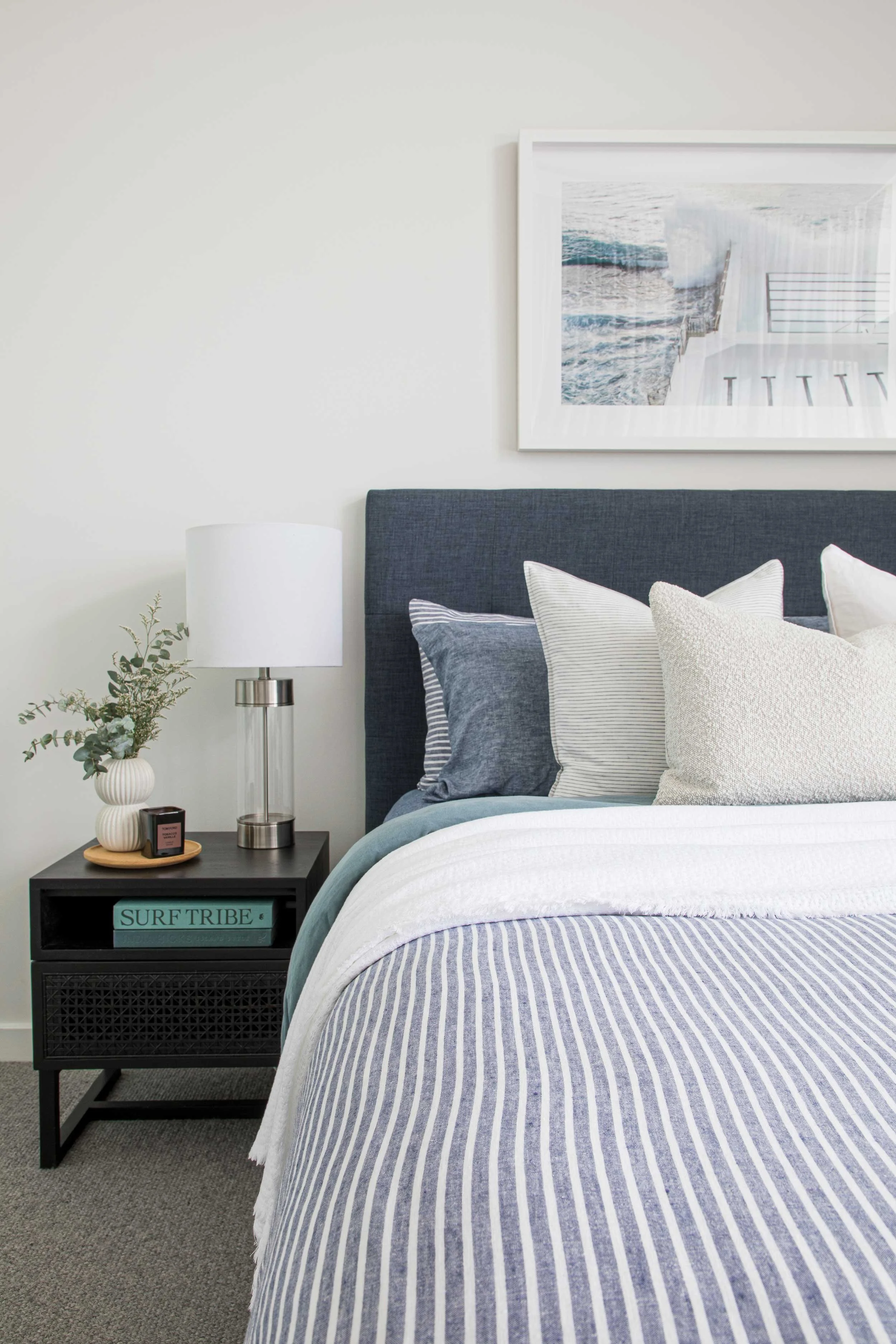 A neatly made bed with striped bedding, multiple pillows, a dark headboard, and a nightstand with a lamp, plant, and books, in a modern bedroom with framed ocean photo on the wall.