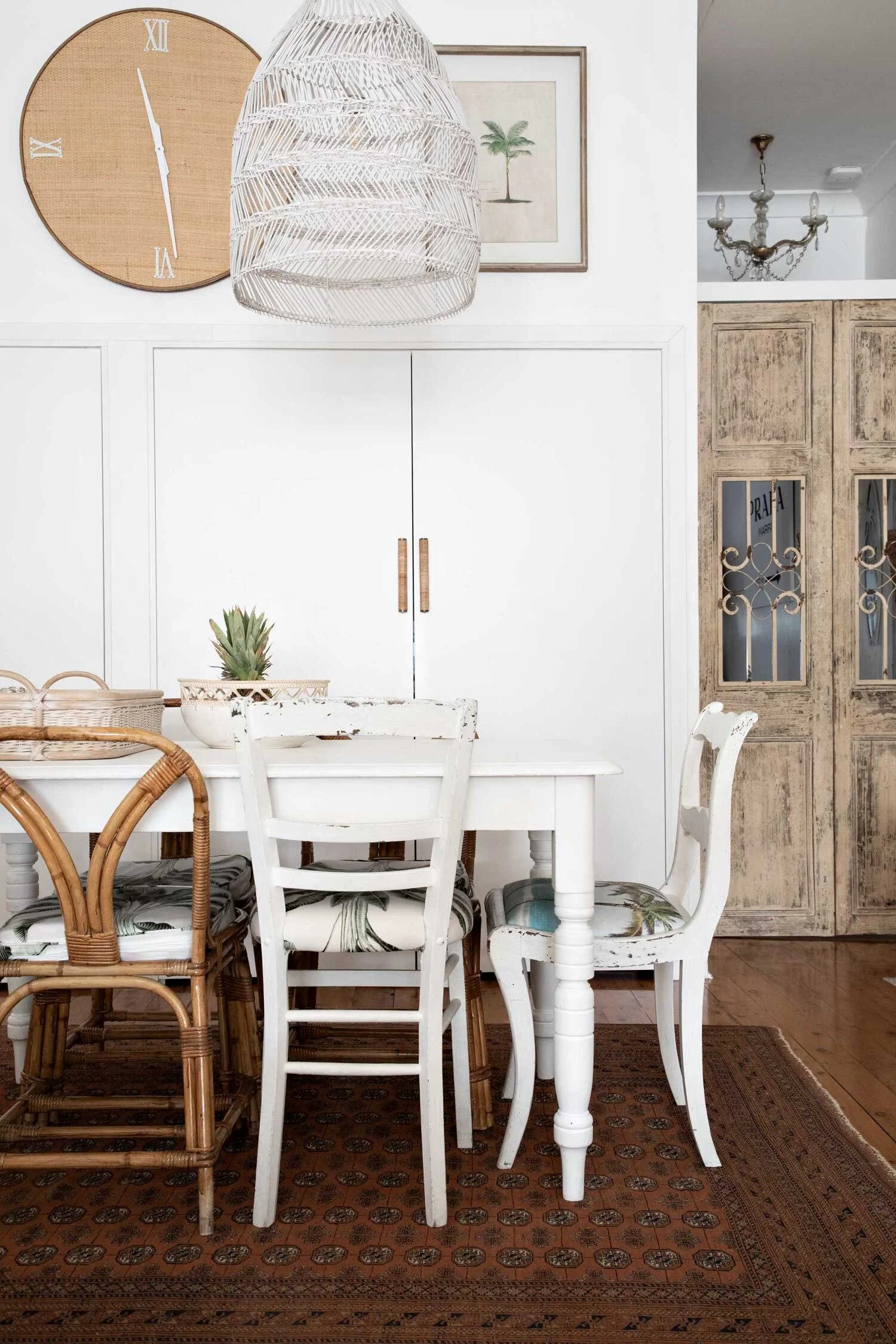 Interior of a dining room with a white table and mixed style chairs, including rattan and painted wood, a pineapple on the table, a patterned rug, a large wall clock, framed artwork, and a woven light fixture, with a rustic cabinet and chandelier in 