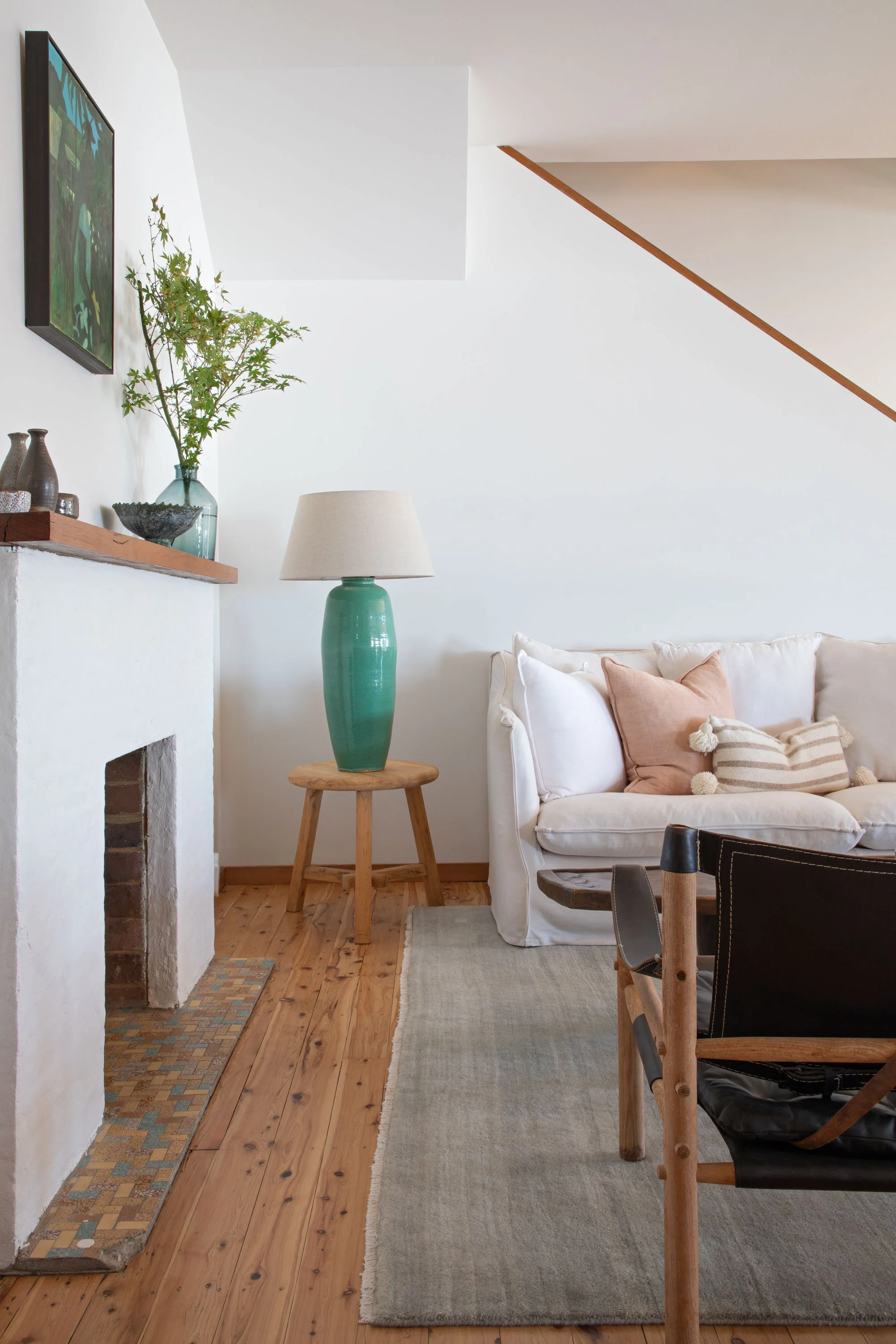 A cozy living room with a white sofa decorated with beige and striped pillows, a large teal ceramic lamp on a small wooden stool, a wooden floor with a small mosaic tile border near a white wall and fireplace, a wall-mounted television, and a narrow 