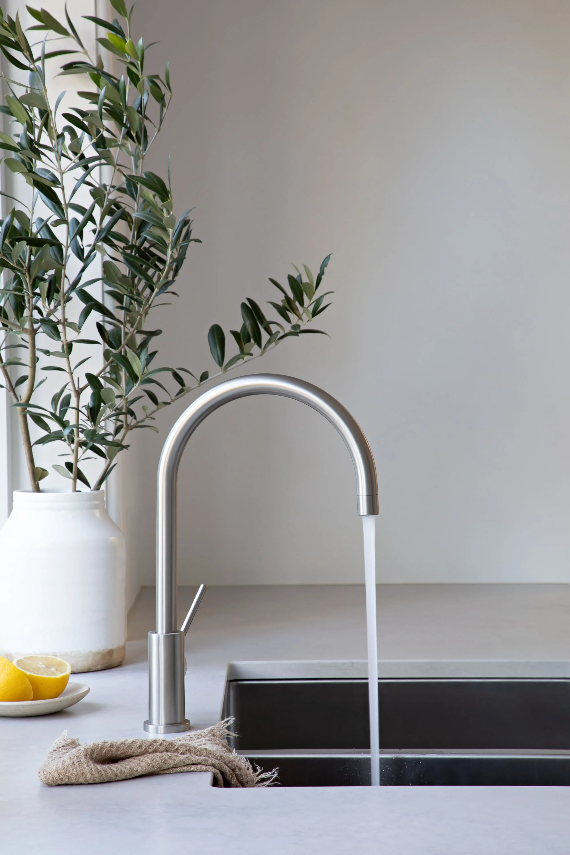 A modern kitchen sink with a stainless steel faucet running water. Behind the sink, there is a white vase with a large green leafy plant. To the left, a small white plate holds two lemon halves. A beige cloth is draped on the countertop in front of t