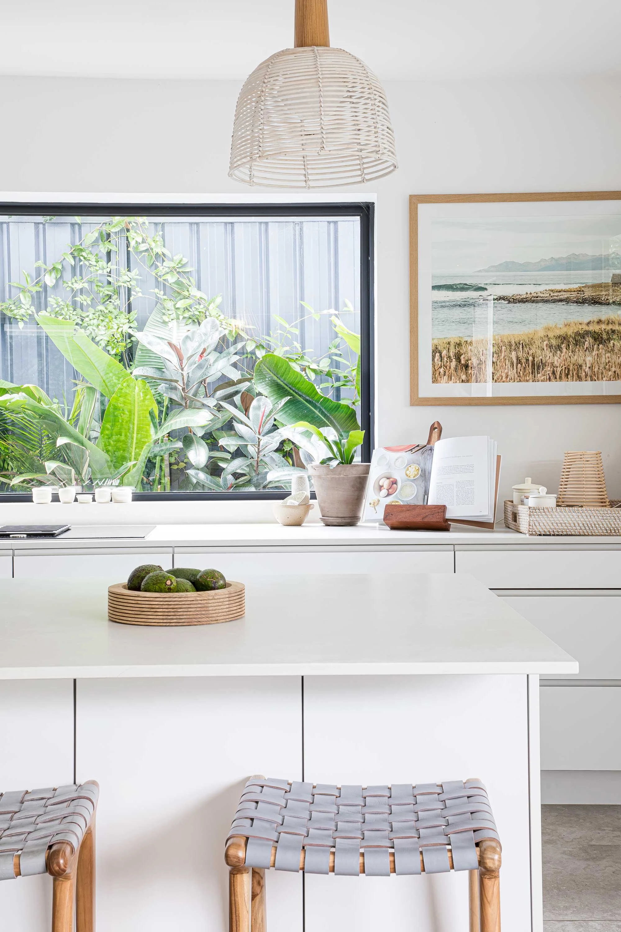 Modern kitchen with white cabinets, a white island, a bowl of avocados, a woven rattan basket, a large framed seaside landscape photo, and green indoor plants visible through a window, with woven stools in the foreground.