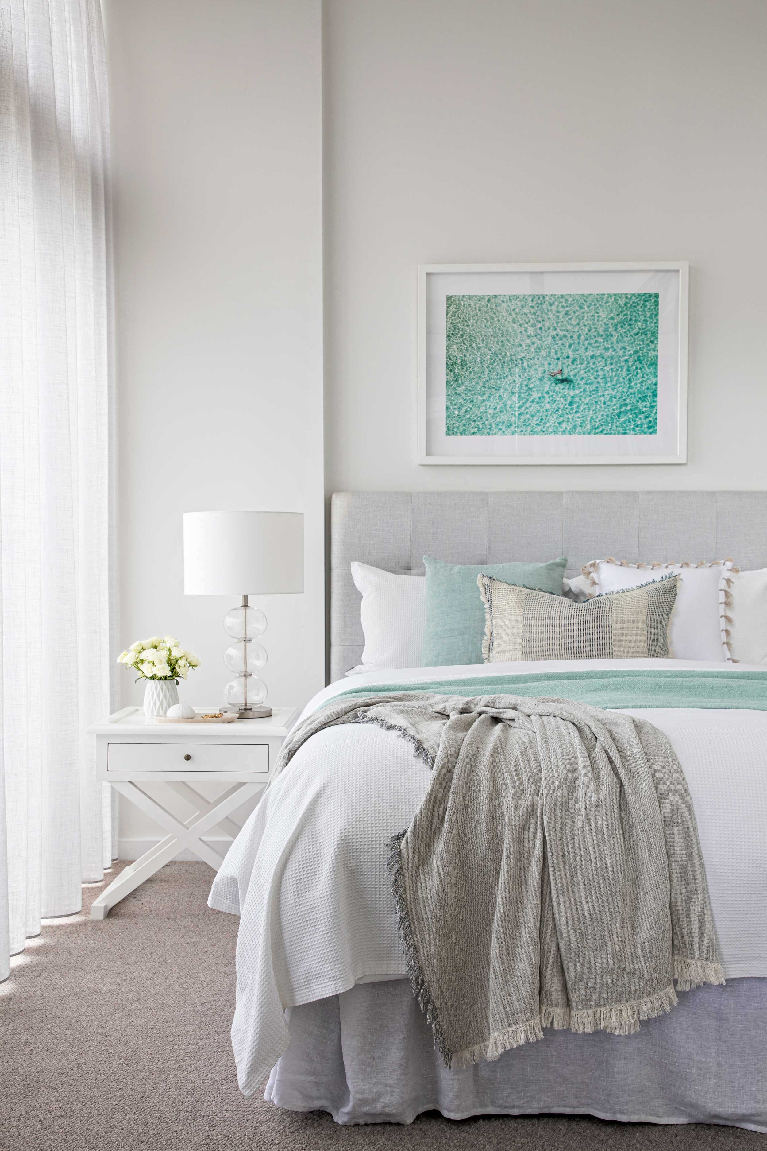 A neatly made bed with white bedding, accented with light green and beige pillows, in a bright bedroom. There is a bedside table with a white lamp, white flowers, and some small items. A framed abstract artwork with turquoise and green tones hangs ab