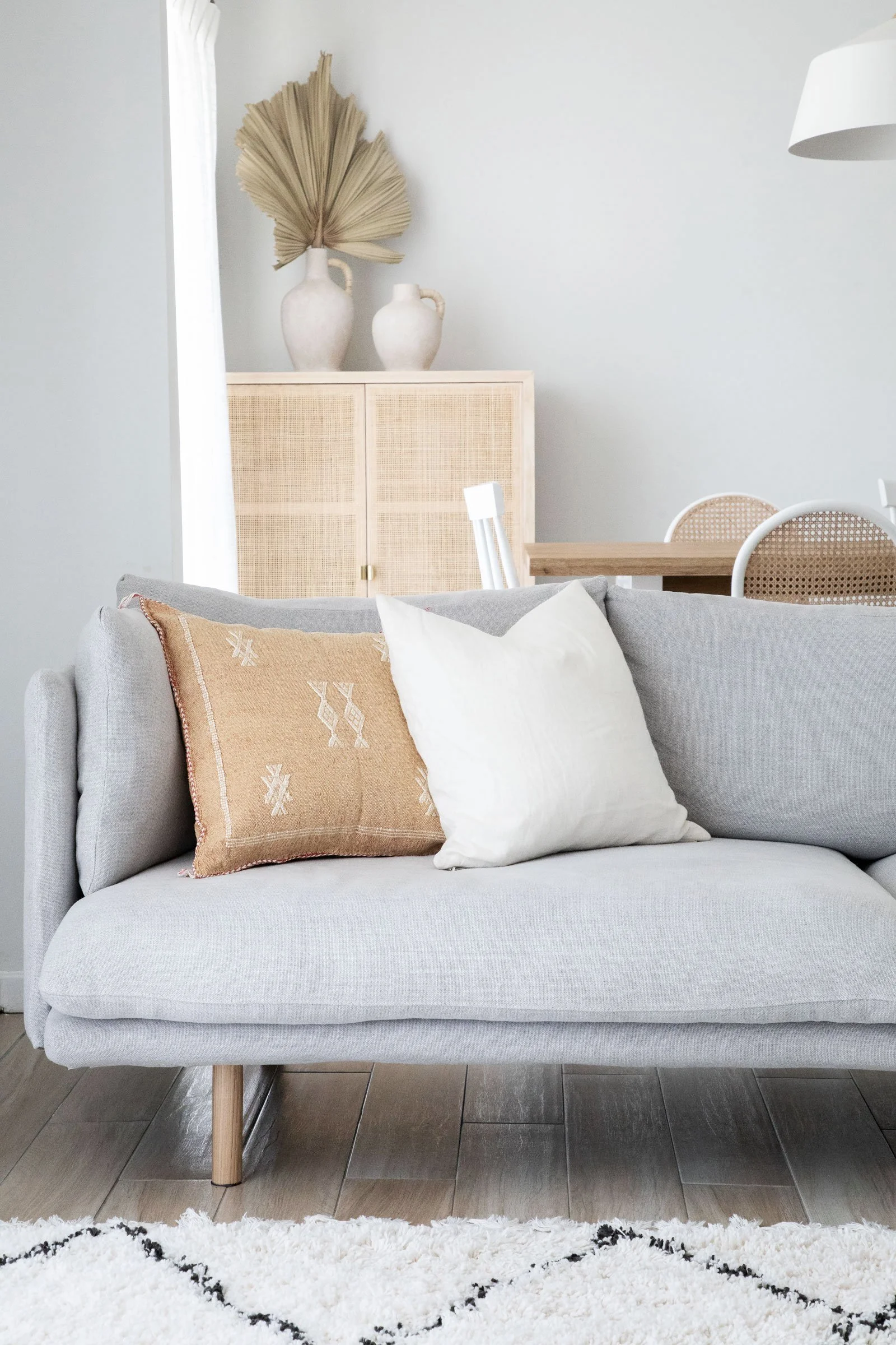 A living room with a light gray sofa that has a beige embroidered pillow and a plain white pillow. Behind the sofa is a wooden table with woven rattan chairs. Against the wall, there is a piece of furniture with two large ceramic vases, one with a dr