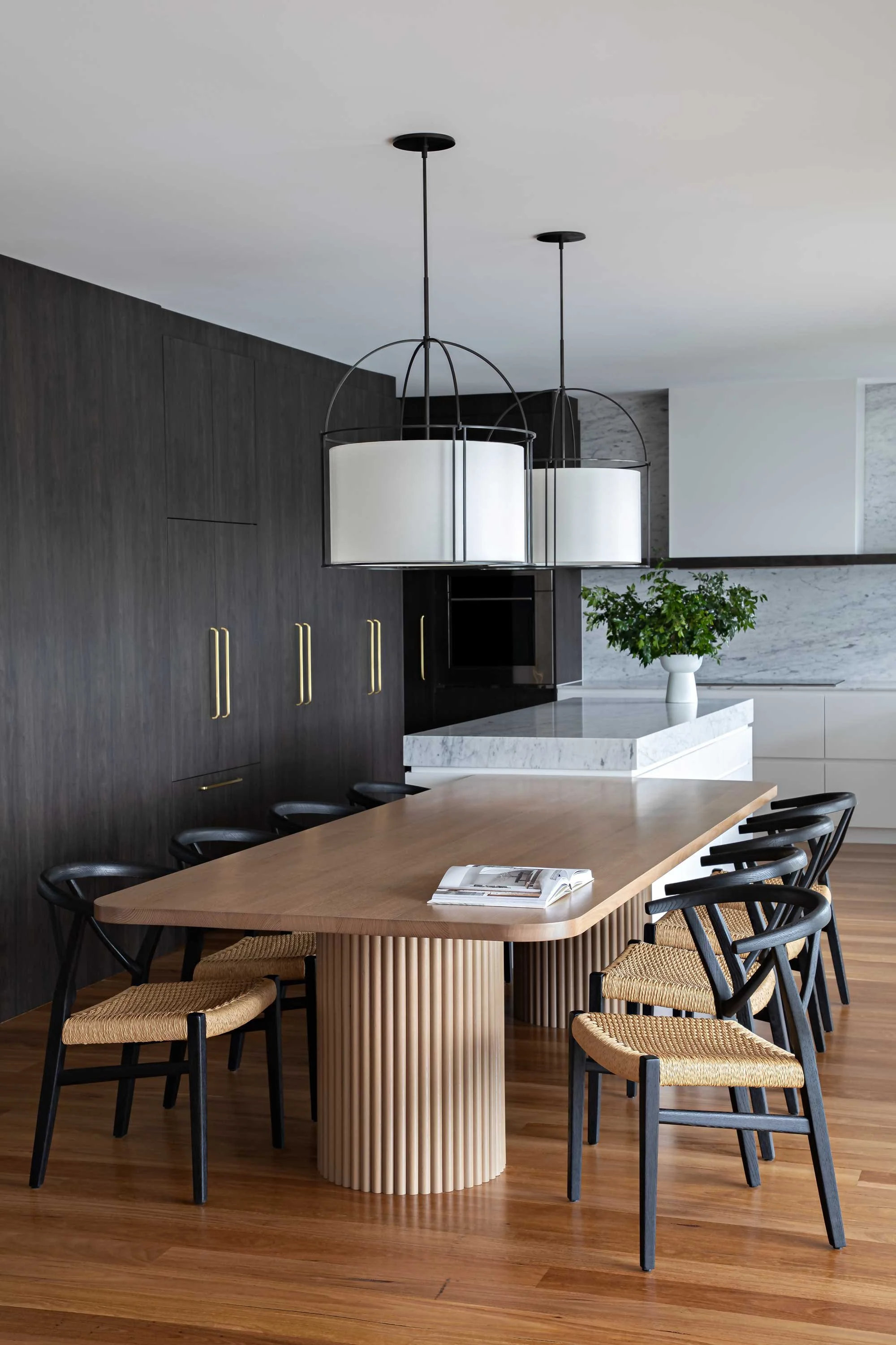 Modern dining room with a wooden table surrounded by black chairs with woven seats, a white marble kitchen island, a dark wood wall with gold handles, a white ceiling, hanging black and white pendant lights, and a white flower pot with green plant on the marble island.