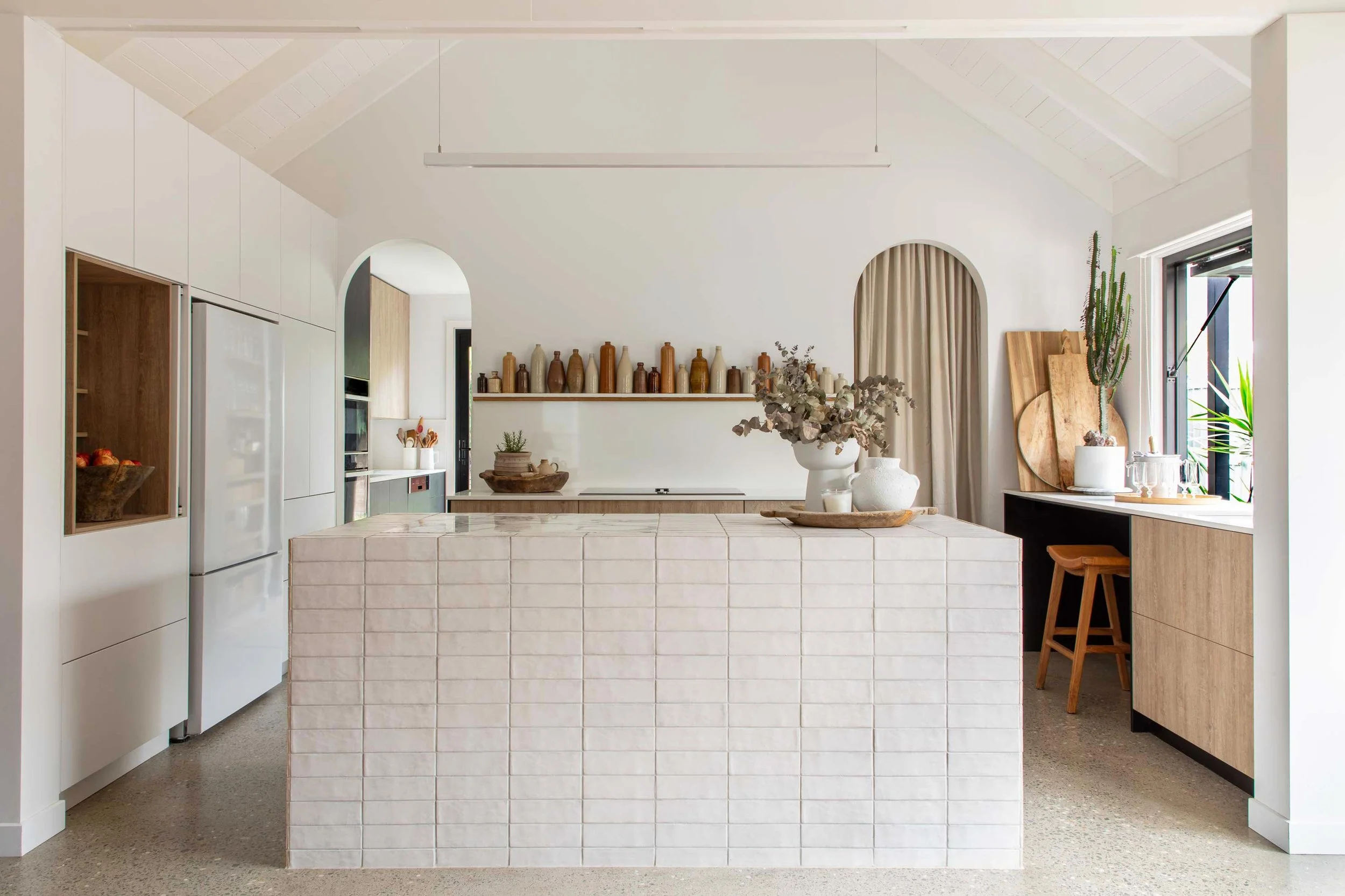 Modern kitchen with white cabinetry, a tiled island in the center, and decorative vases on a shelf above the stove. Natural light from a window on the right illuminates plants and wooden accents.