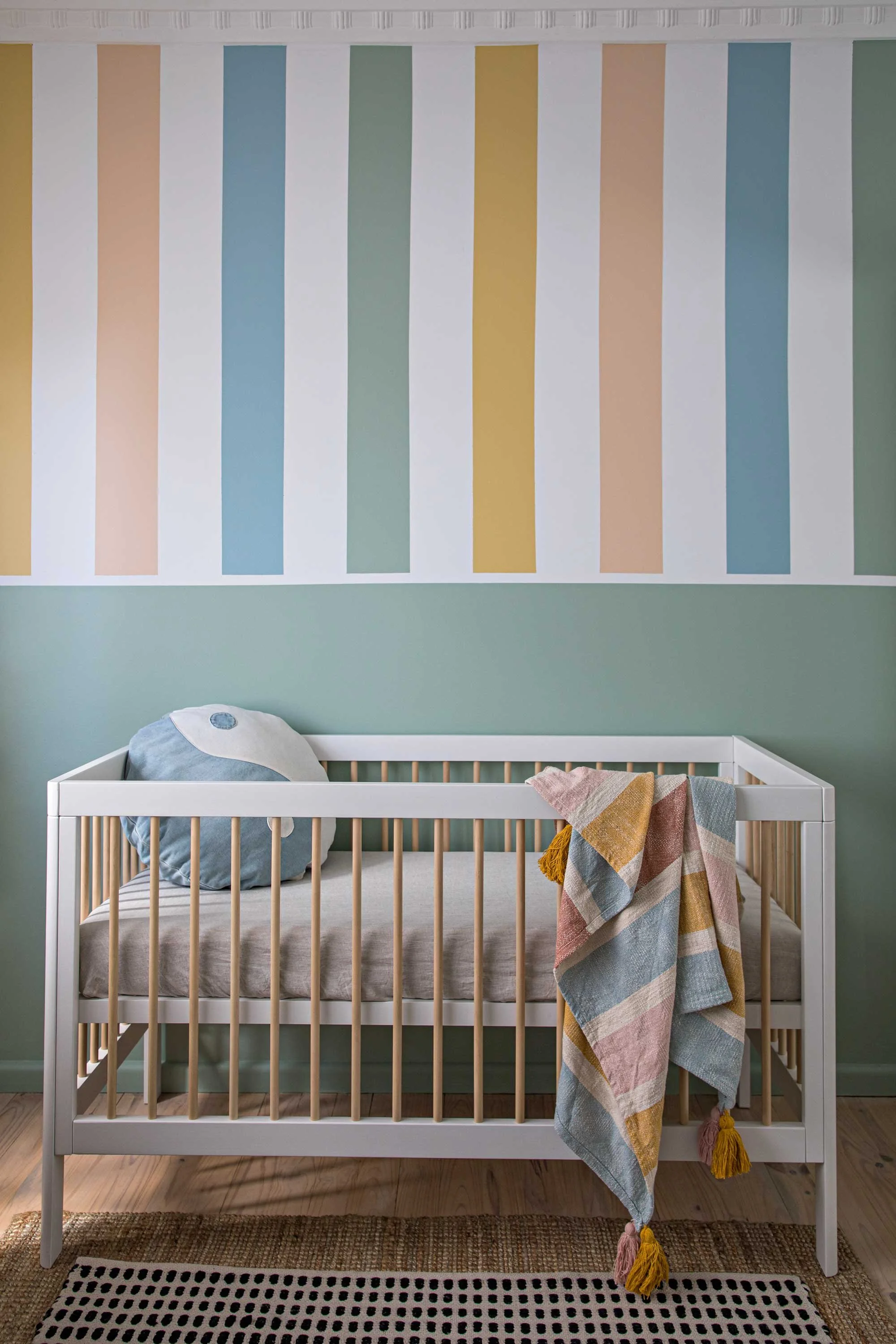 A white crib with beige mattress and wooden slats, situated against a pastel-colored wall with vertical stripes in shades of green, blue, peach, and yellow. A crumpled, multicolored blanket with tassels hangs over the side of the crib. A grey plush p