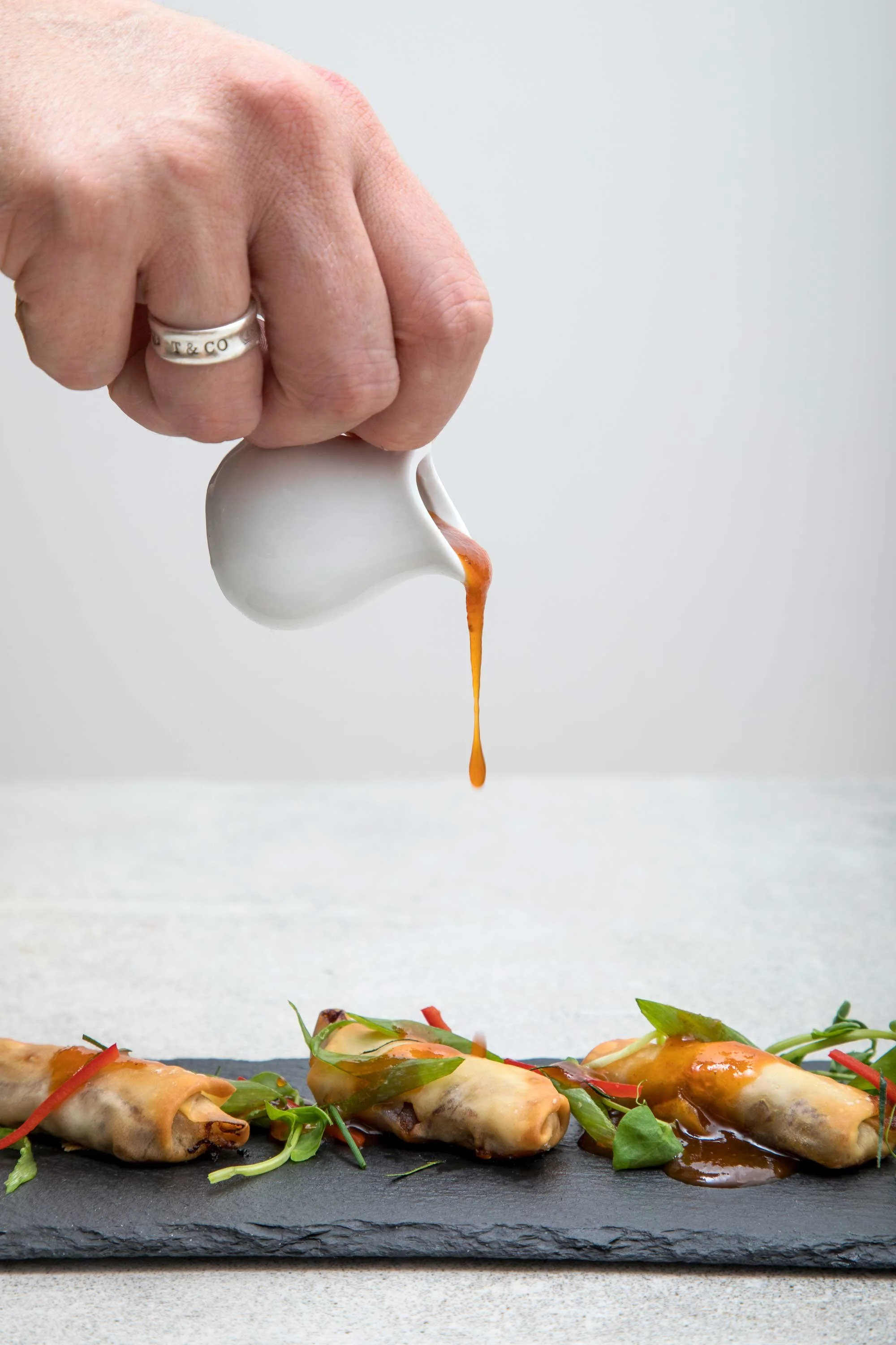 A hand pouring sauce over a row of spring rolls garnished with greens on a black slate platter.