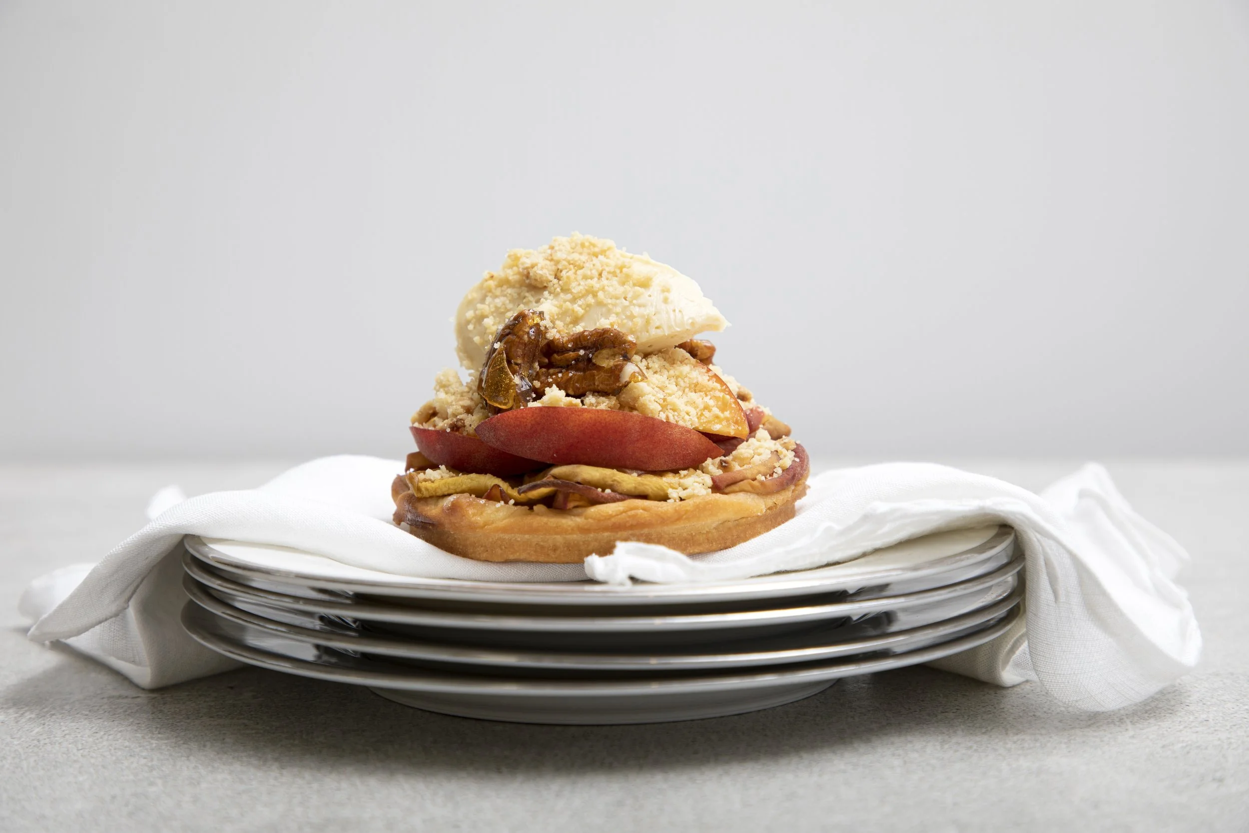 A stack of freshly baked fruit tart with slices of red apple, caramelized pecans, crumbled topping, a scoop of vanilla ice cream, and crumbs on top, placed on a white cloth and set on stacked white plates.