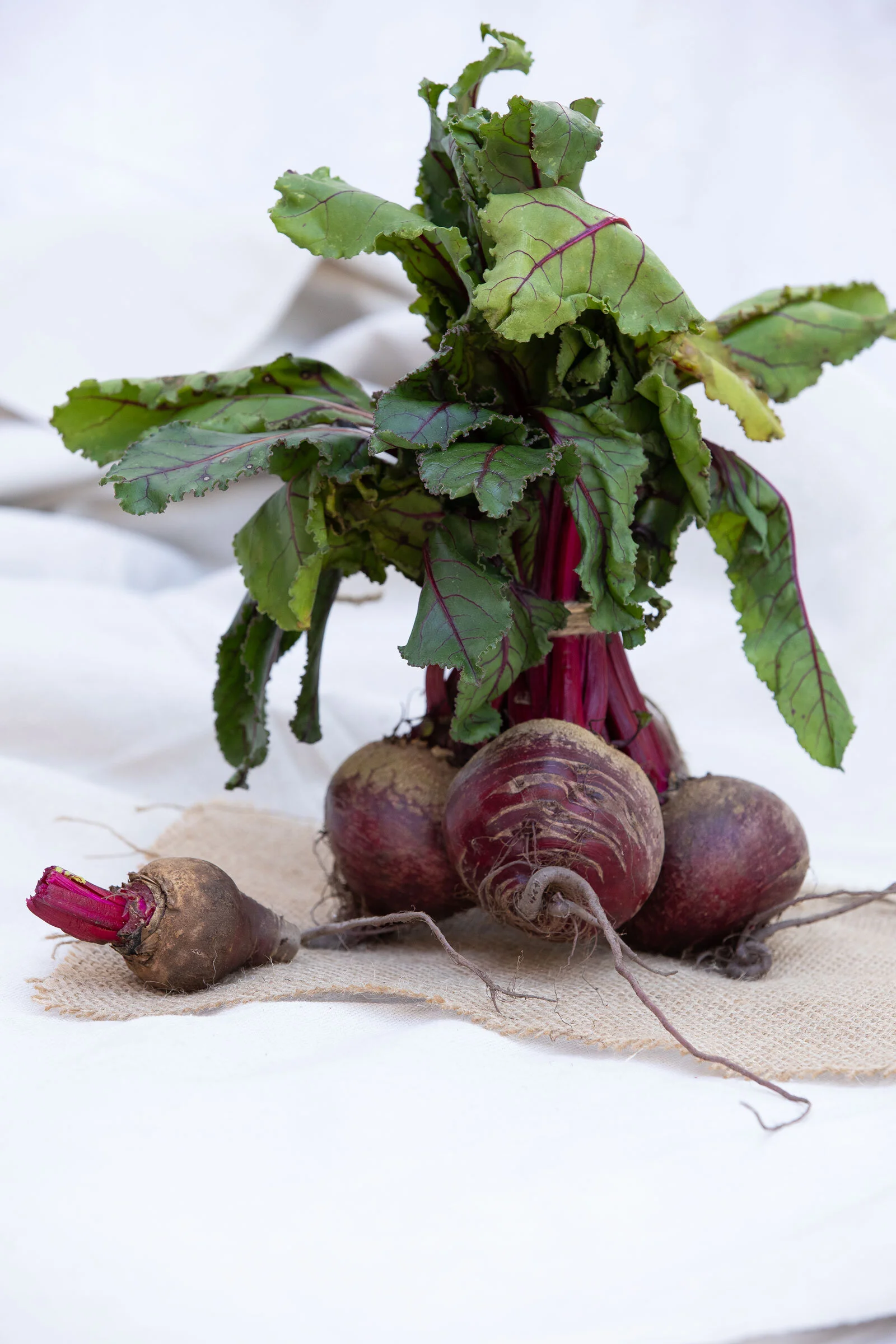 Fresh beetroots with green leaves, some with visible red stems, resting on a light cloth surface.