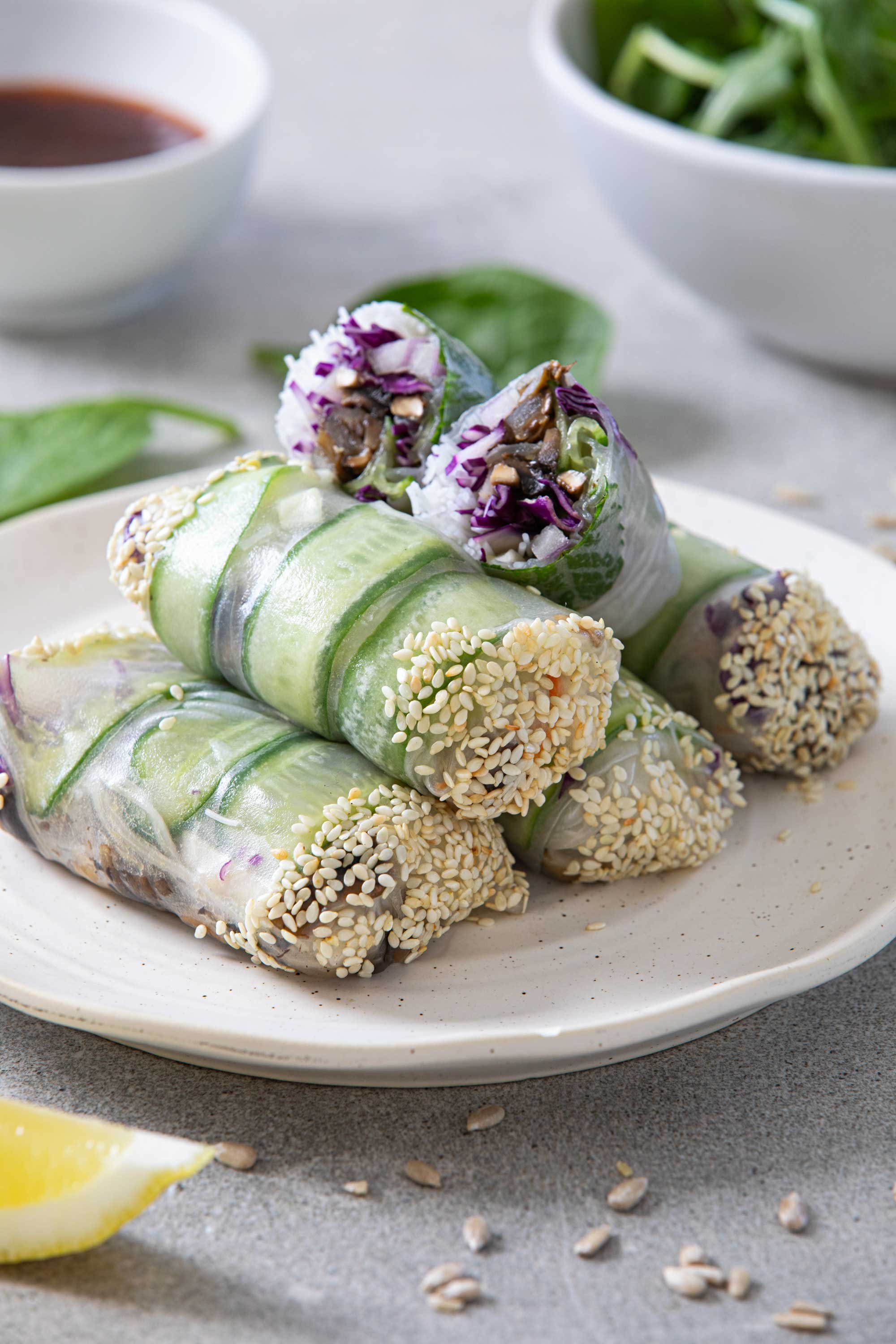 Close-up of fresh spring rolls filled with vegetables and herbs, garnished with sesame seeds, served on a white plate with a lemon wedge and sesame seeds scattered around.