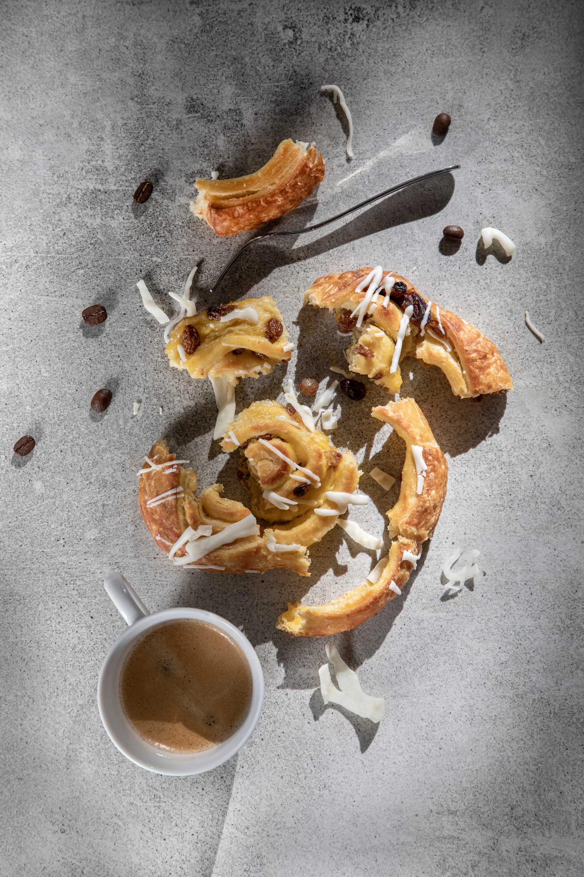 Broken pastry with white icing and chocolate chips on a grey surface, a small cup of coffee nearby, scattered pastry remnants and chocolate chips.