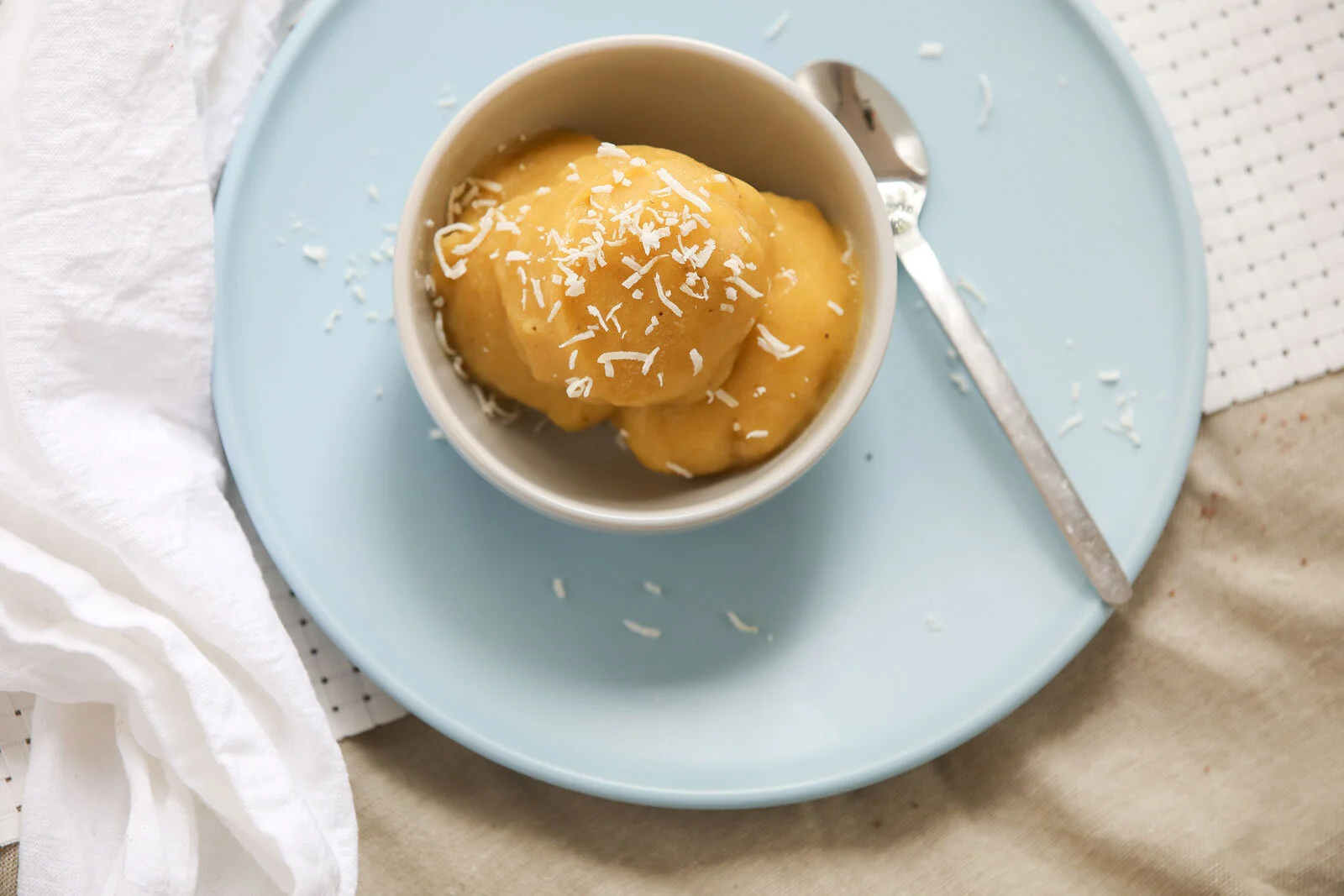 A small white bowl containing three scoops of yellow ice cream with shredded white chocolate on top, placed on a light blue plate with a silver spoon nearby. The setting includes a white napkin and a beige textured surface.