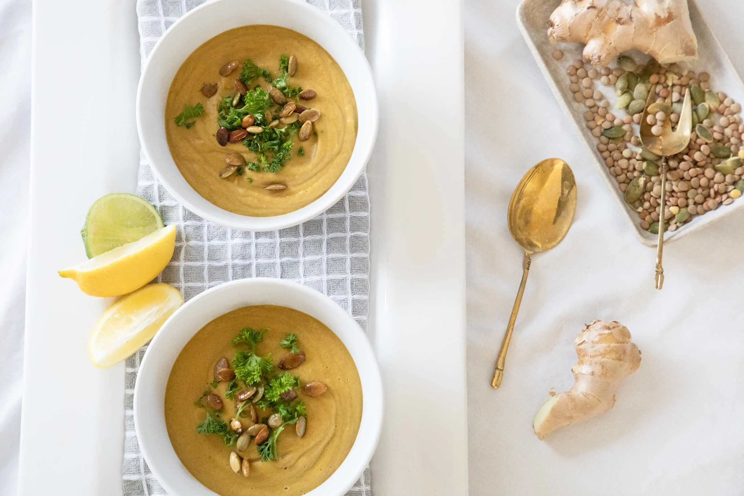 Two bowls of creamy soup garnished with cilantro and pumpkin seeds, lemon wedges on the side, and ginger roots and lentils on a white surface.