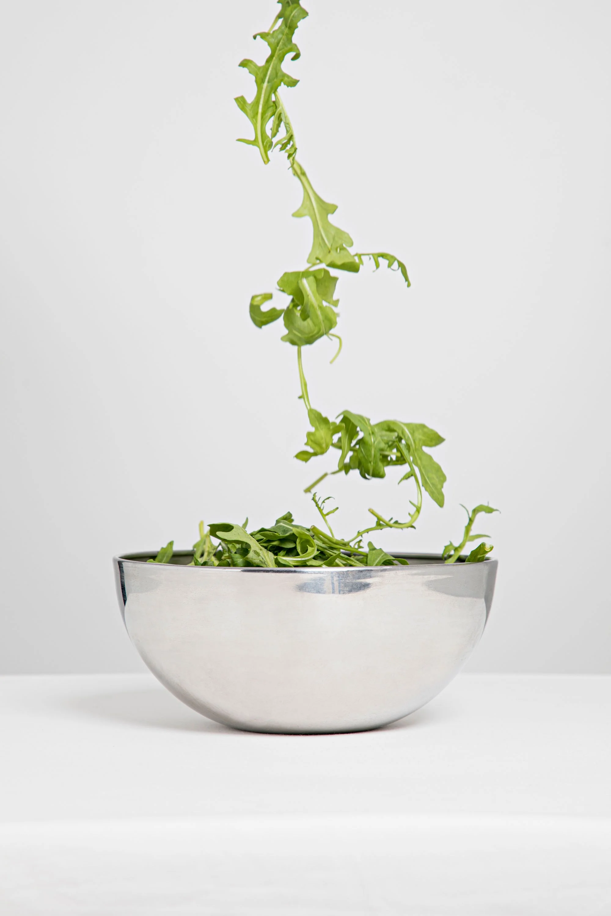 Fresh green leafy arugula being dropped into a white ceramic bowl against a plain white background.