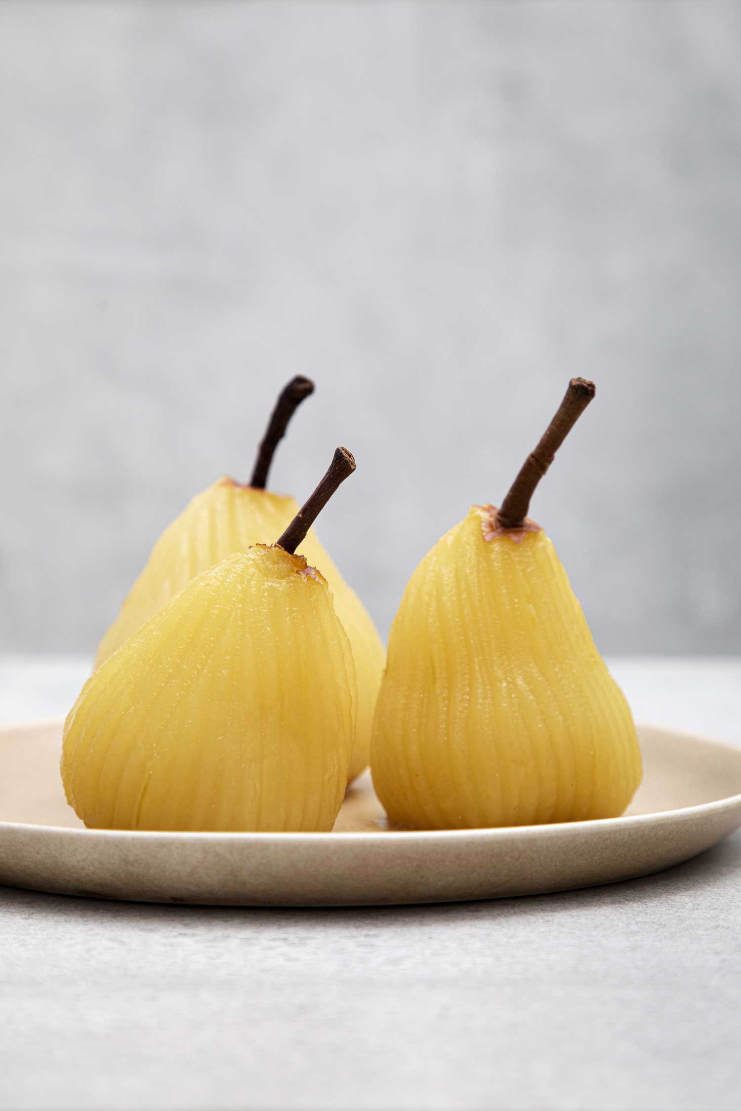Three peeled yellow pears with stems on a white plate against a gray background.
