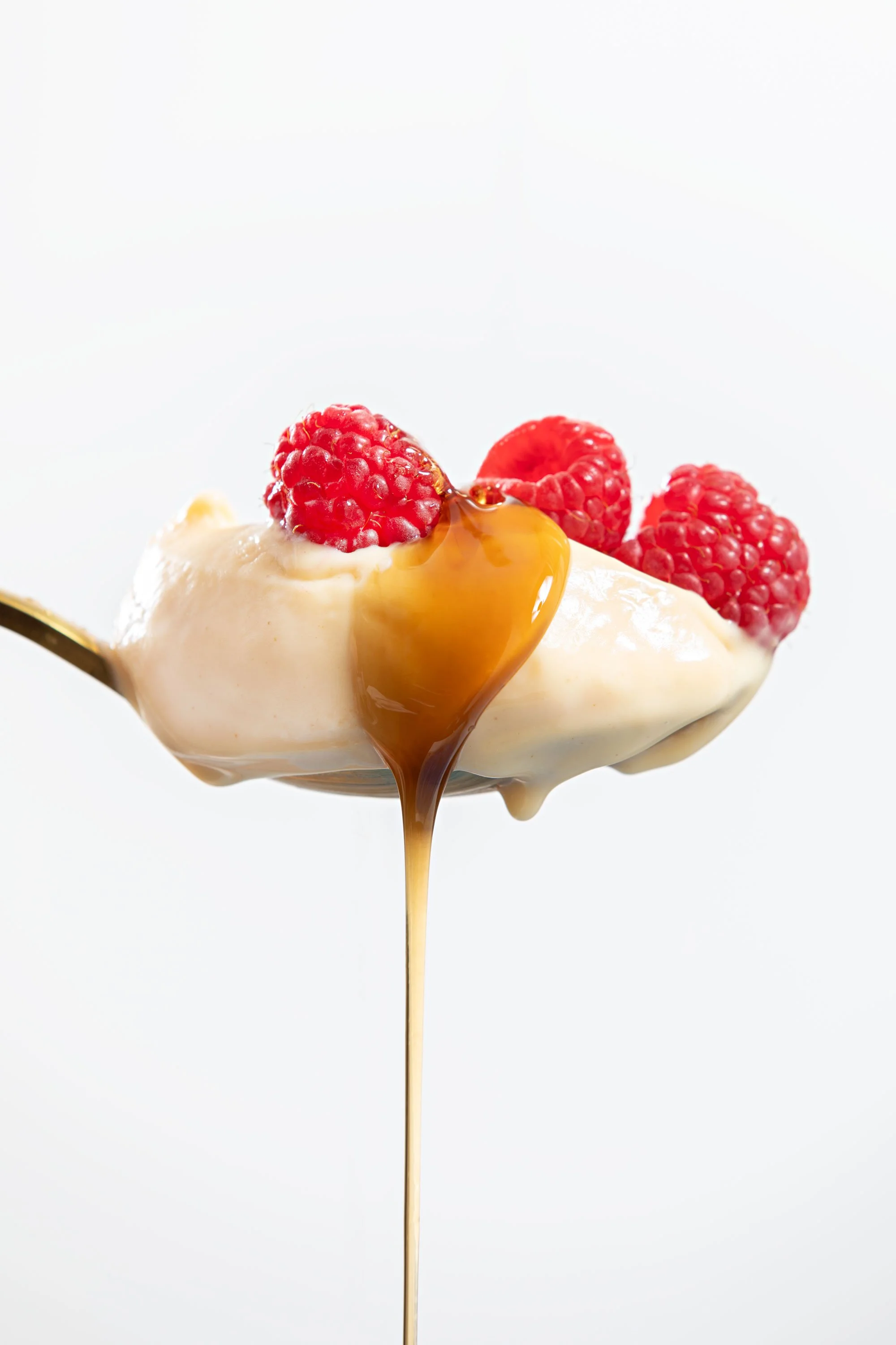 Close-up of a spoonful of creamy dessert topped with raspberries and drizzled with caramel sauce, against a white background.