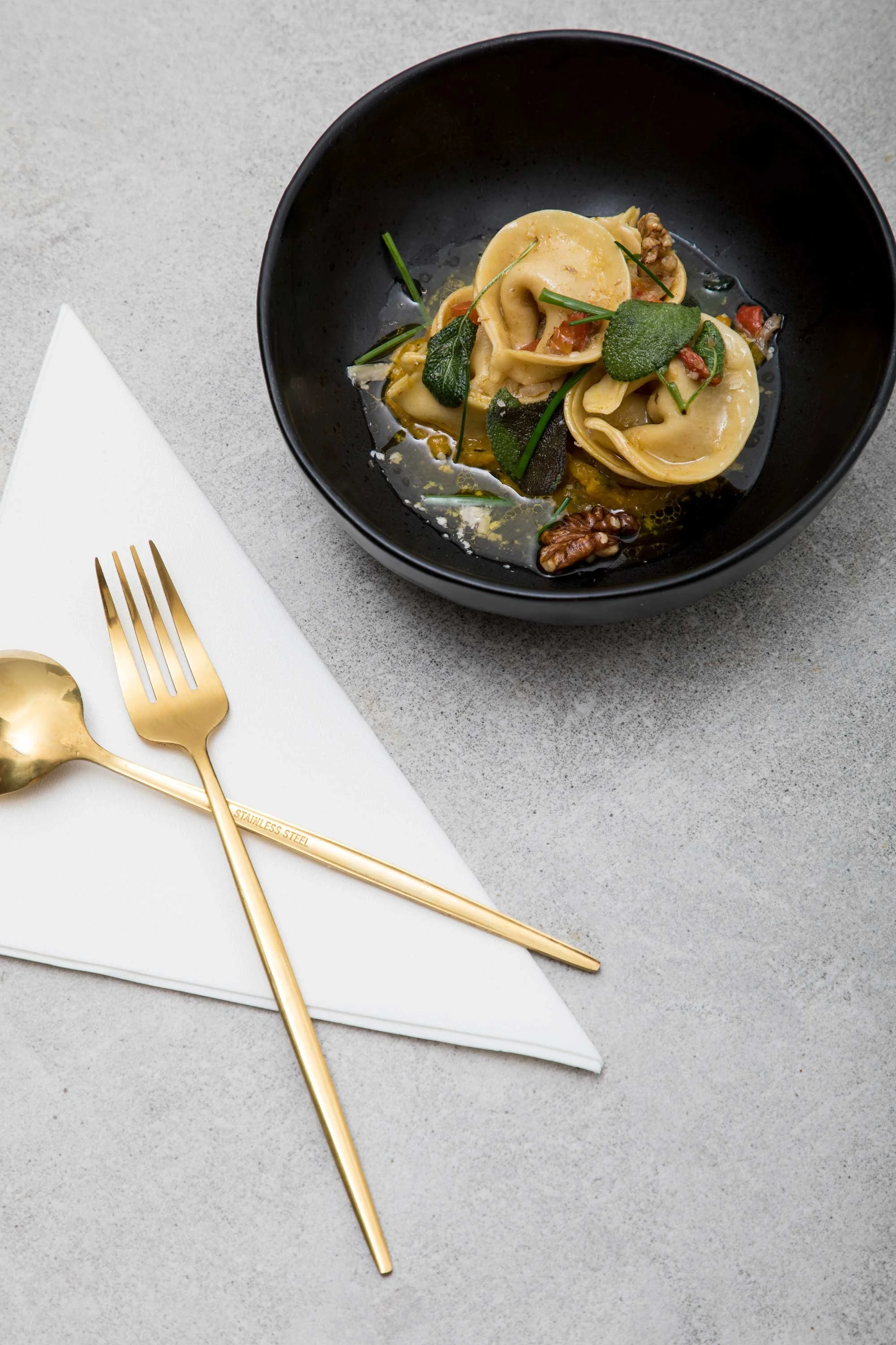 A black bowl containing cooked ravioli pasta with dark greens, walnuts, and a light sauce on a gray surface.