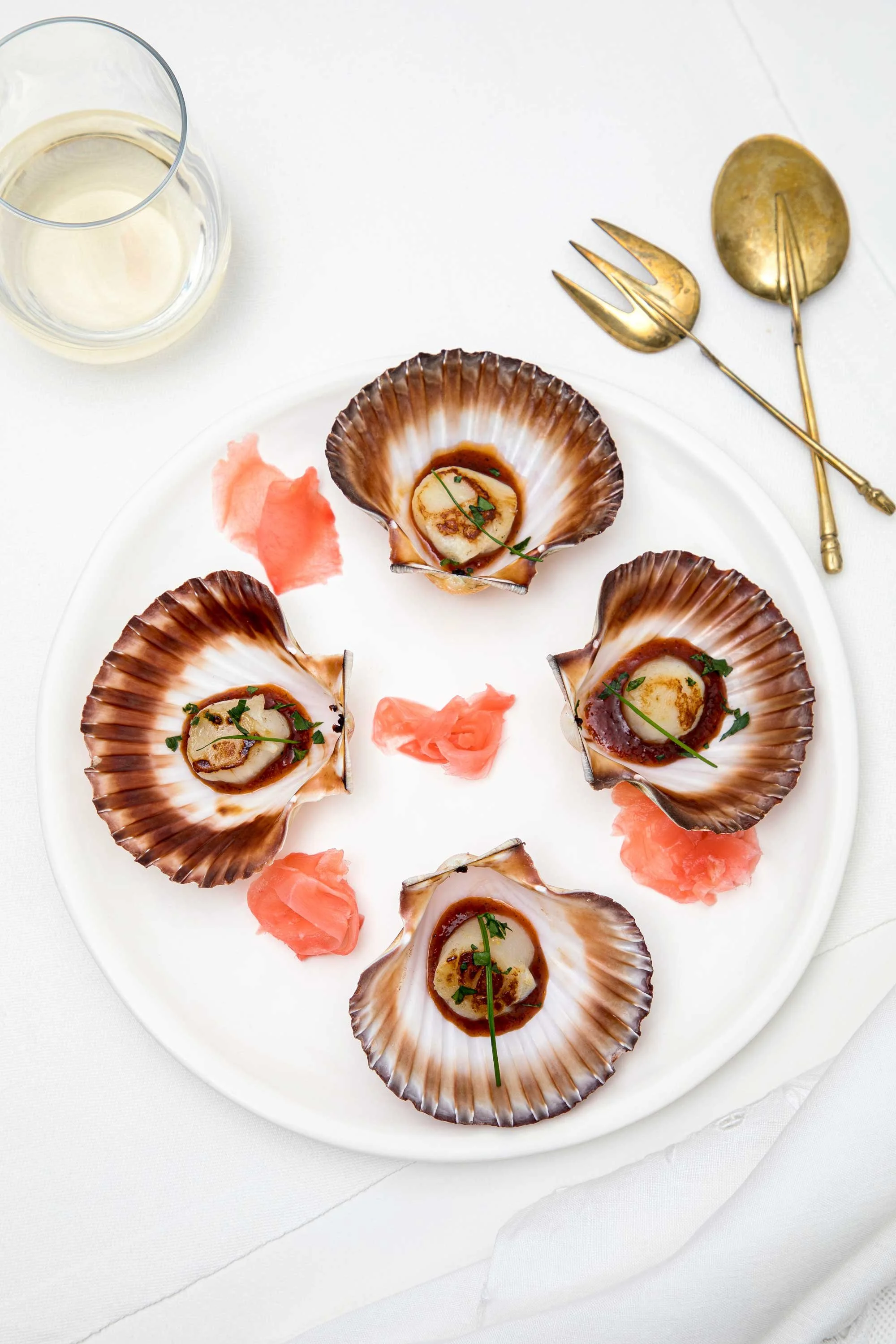 A white plate with several cooked scallops in their shells, garnished with herbs and sauce, with pink pickled ginger around them. A glass of white wine and gold-colored serving utensils are visible on a white tablecloth.
