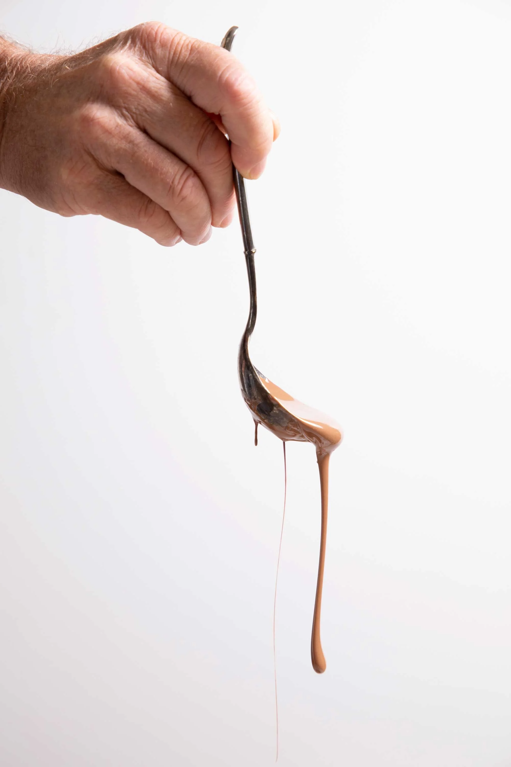 Close-up of a hand holding a spoon with melted chocolate dripping from it, against a plain white background.