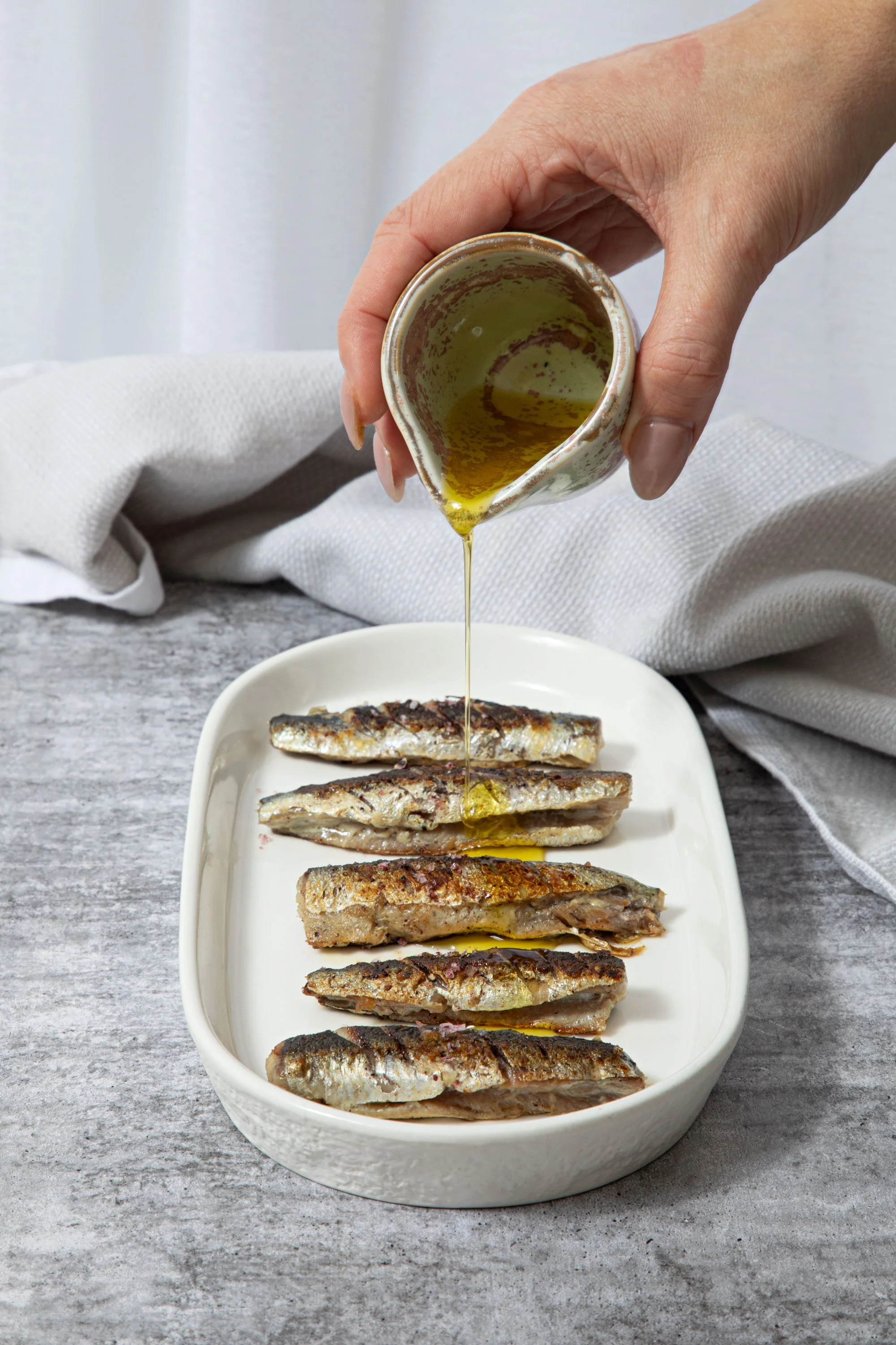 A hand pouring olive oil over grilled fish fillets on a white oval dish. The background is a light wooden surface with a white cloth and a gray textured table.