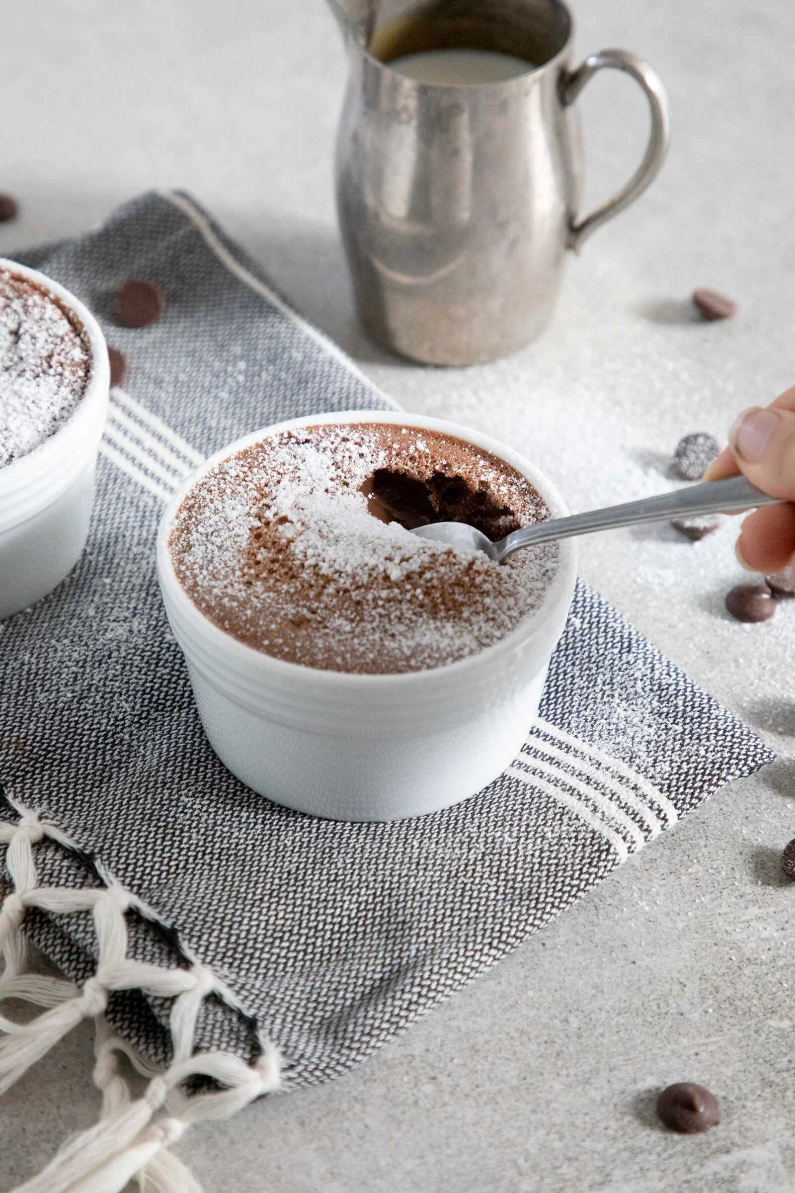 A ceramic cup of hot chocolate topped with powdered sugar, resting on a woven gray cloth with a striped pattern, with a metallic pitcher of milk and scattered chocolate candies in the background. A hand is holding a spoon in the hot chocolate.