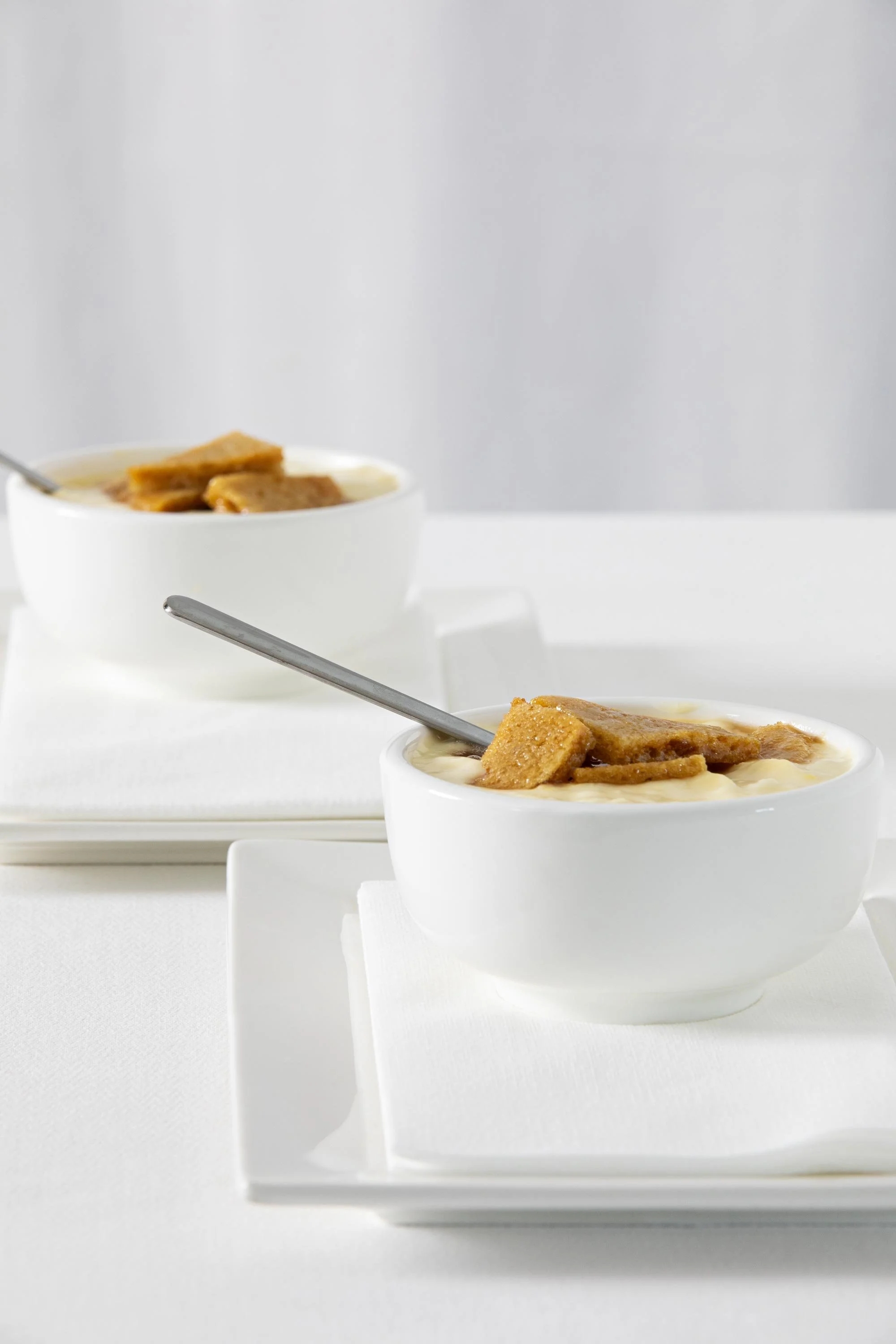 Two white bowls of bread pudding with gold-brown bread chunks on top, served with a spoon, on white trays against a light gray background.