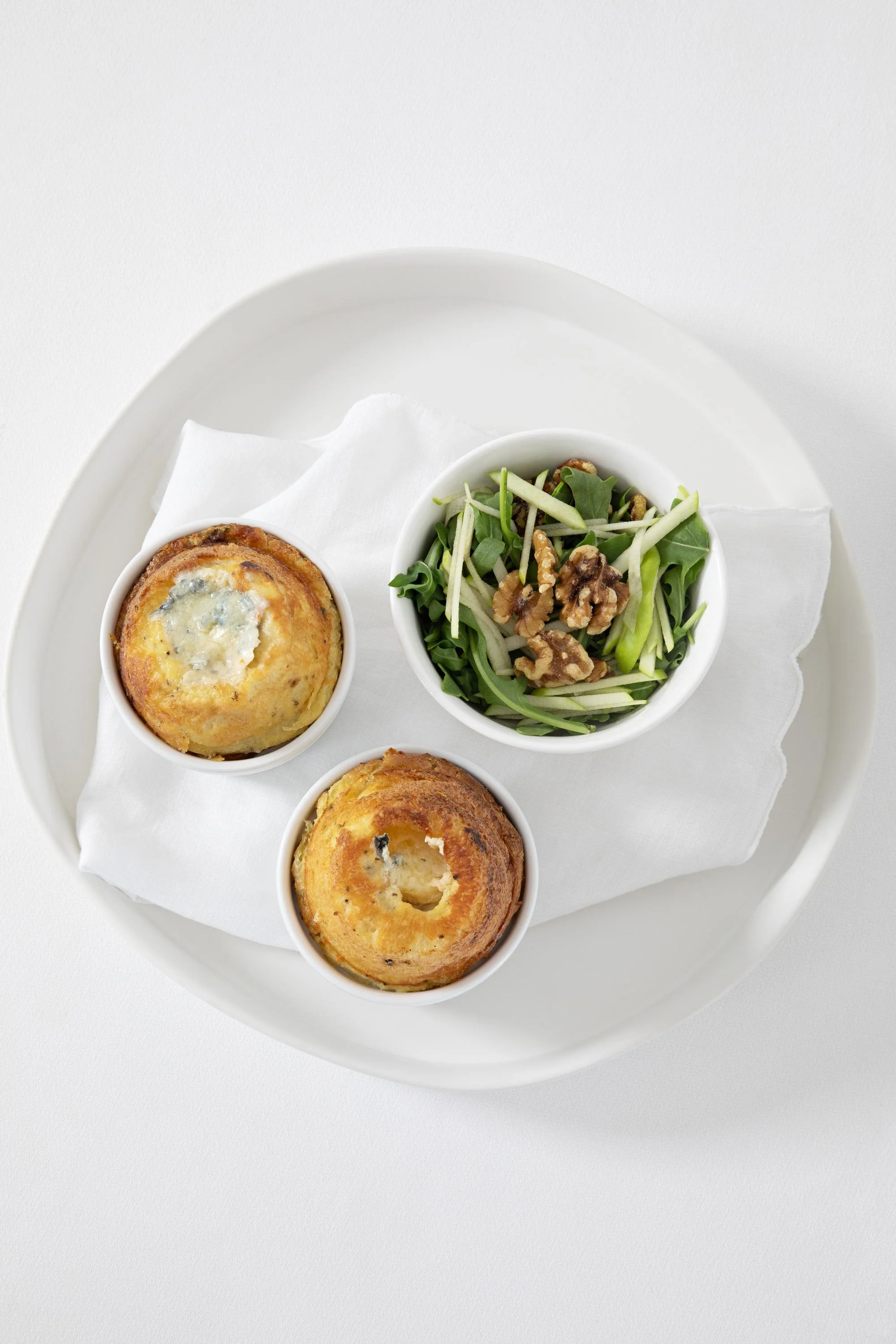Three small ramekins, containing two cheese-filled biscuits and a side salad with walnuts, spinach, and shredded vegetables, arranged on a white tray with a paper napkin.