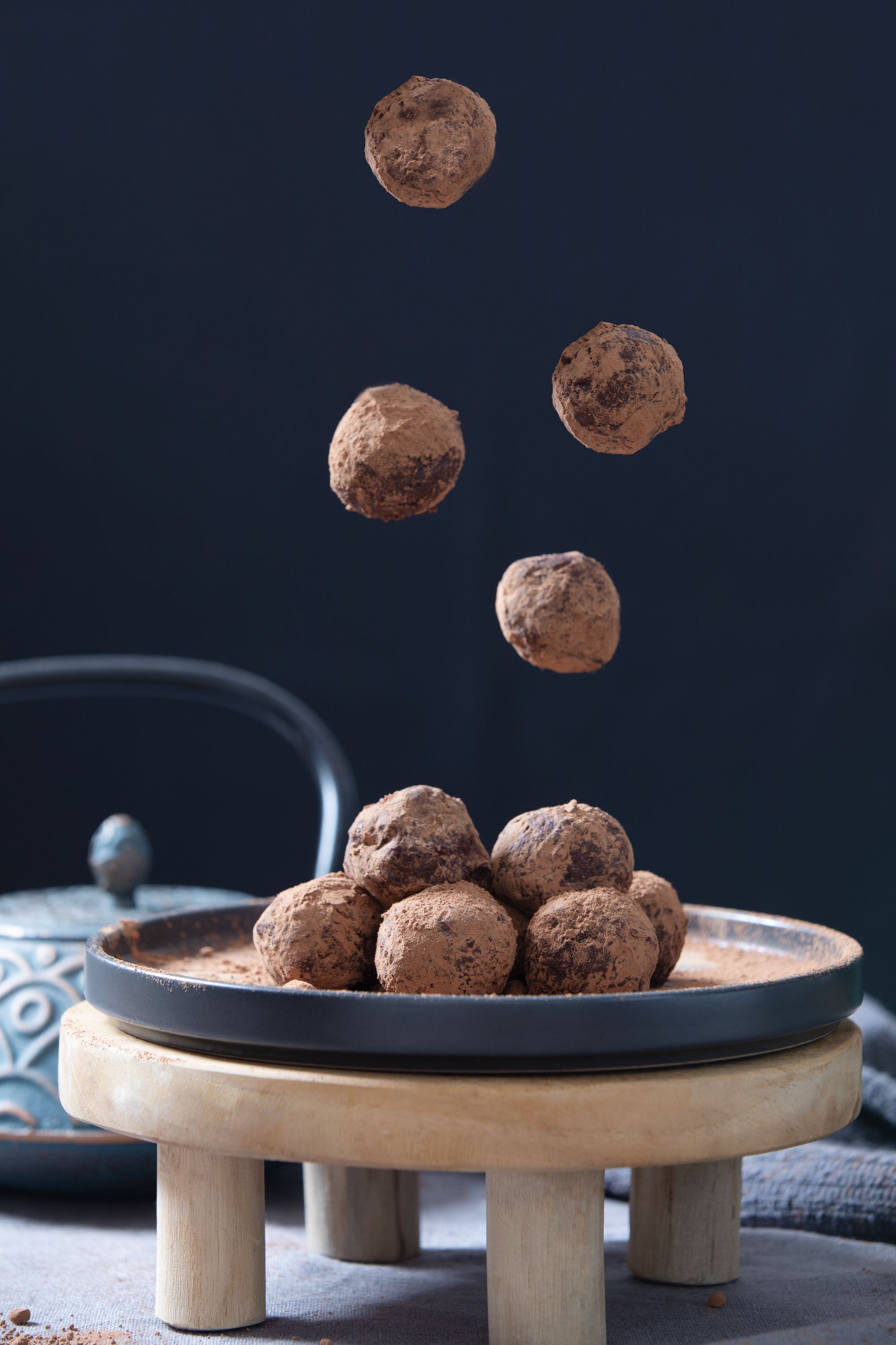 A small wooden stand with a plate of chocolate truffles being dropped into the air, dusted with cocoa powder, against a dark background.
