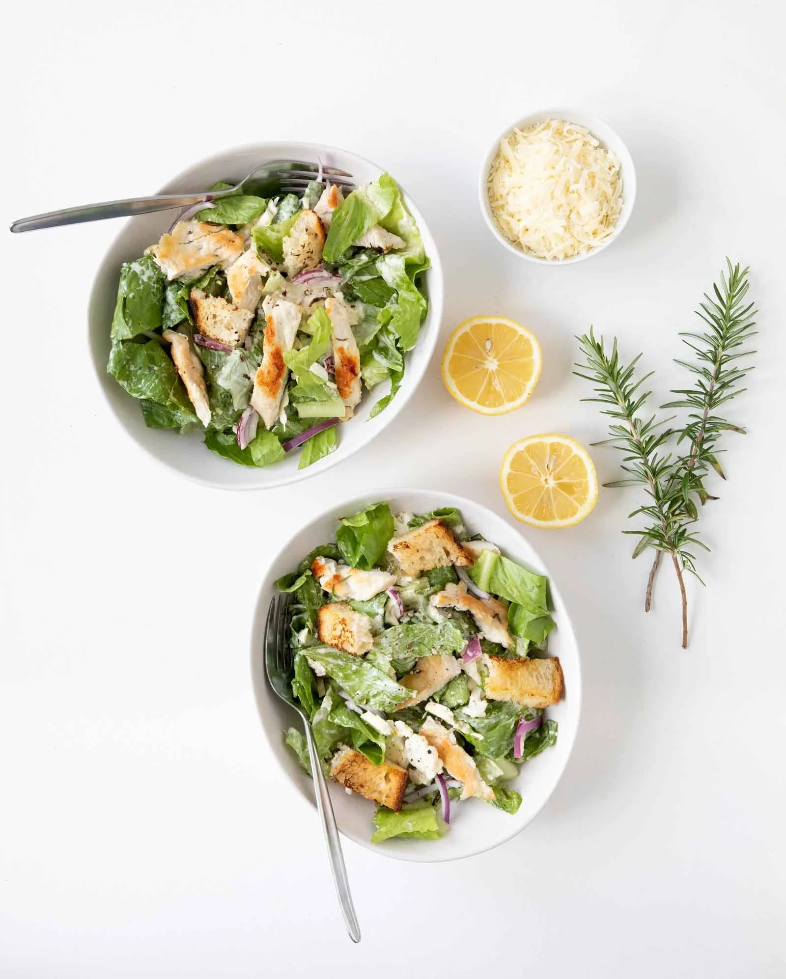 Two bowls of Caesar salad topped with grilled chicken, grated cheese, and croutons, with lemon halves and sprigs of rosemary on white background.