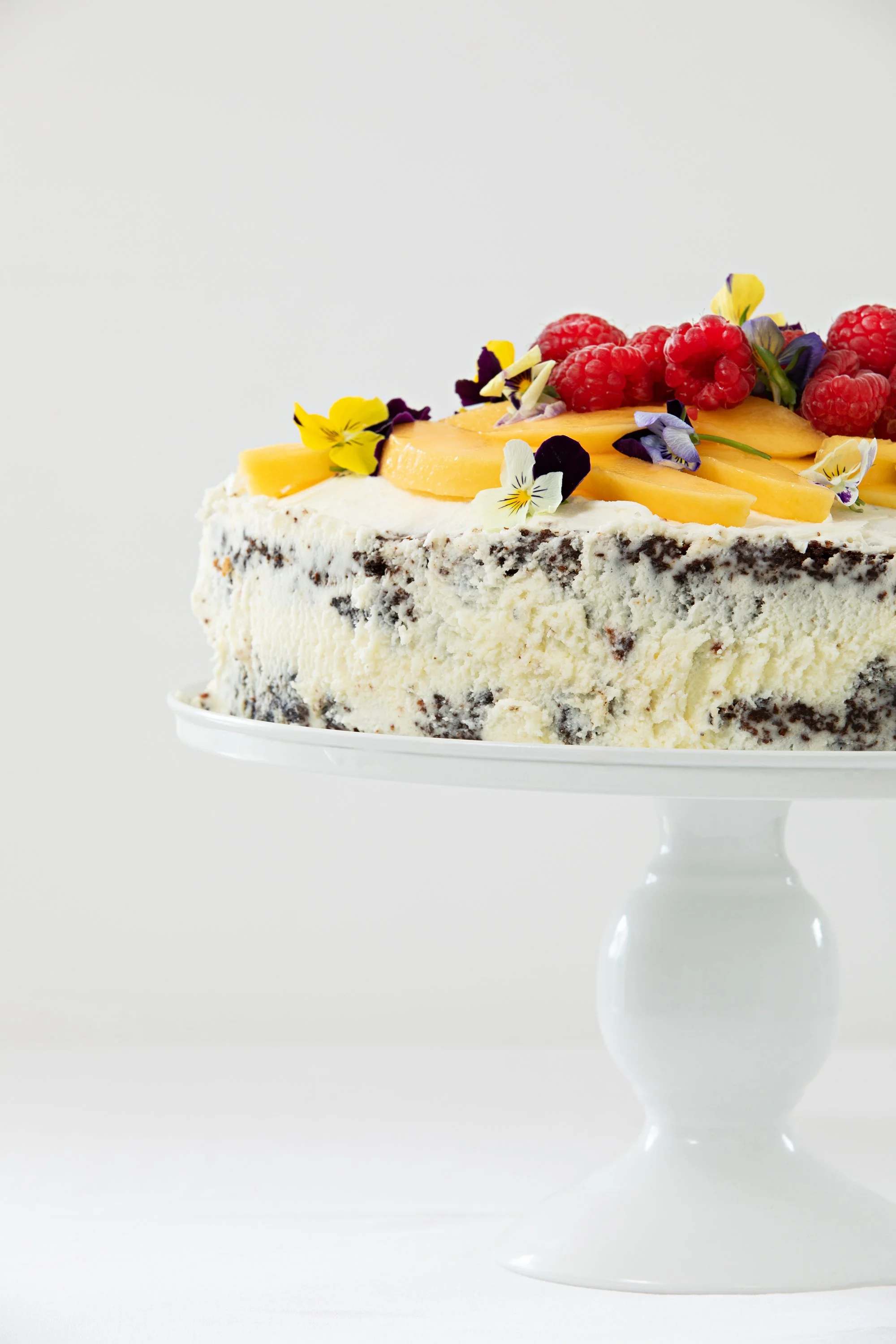 Chocolate and vanilla layered cake topped with raspberries, sliced peaches, and edible flowers on a white cake stand.