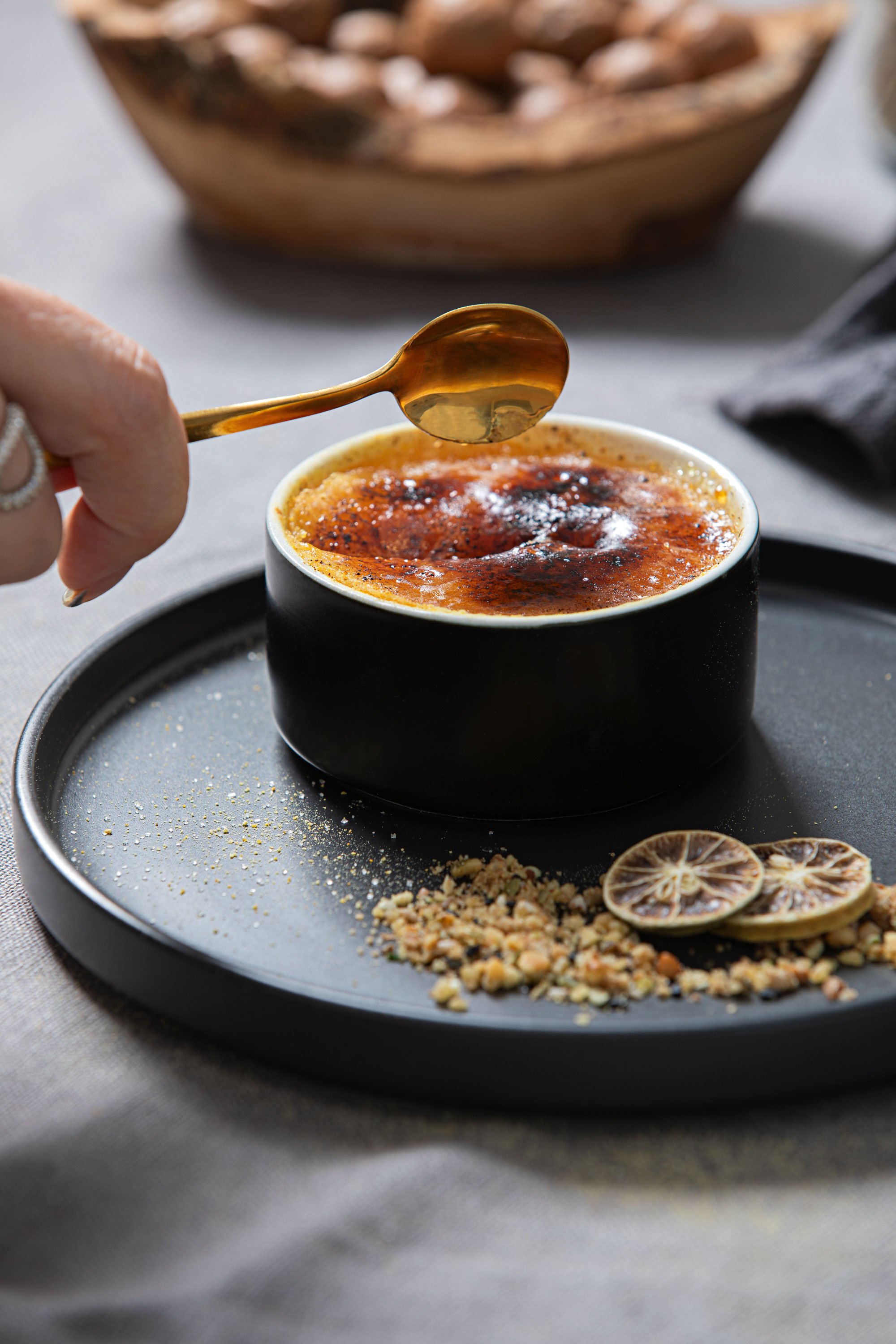 A black cup with a crème brulee dessert topped with caramelized sugar, beside slices of dried lemon and crushed nuts on a black tray.
