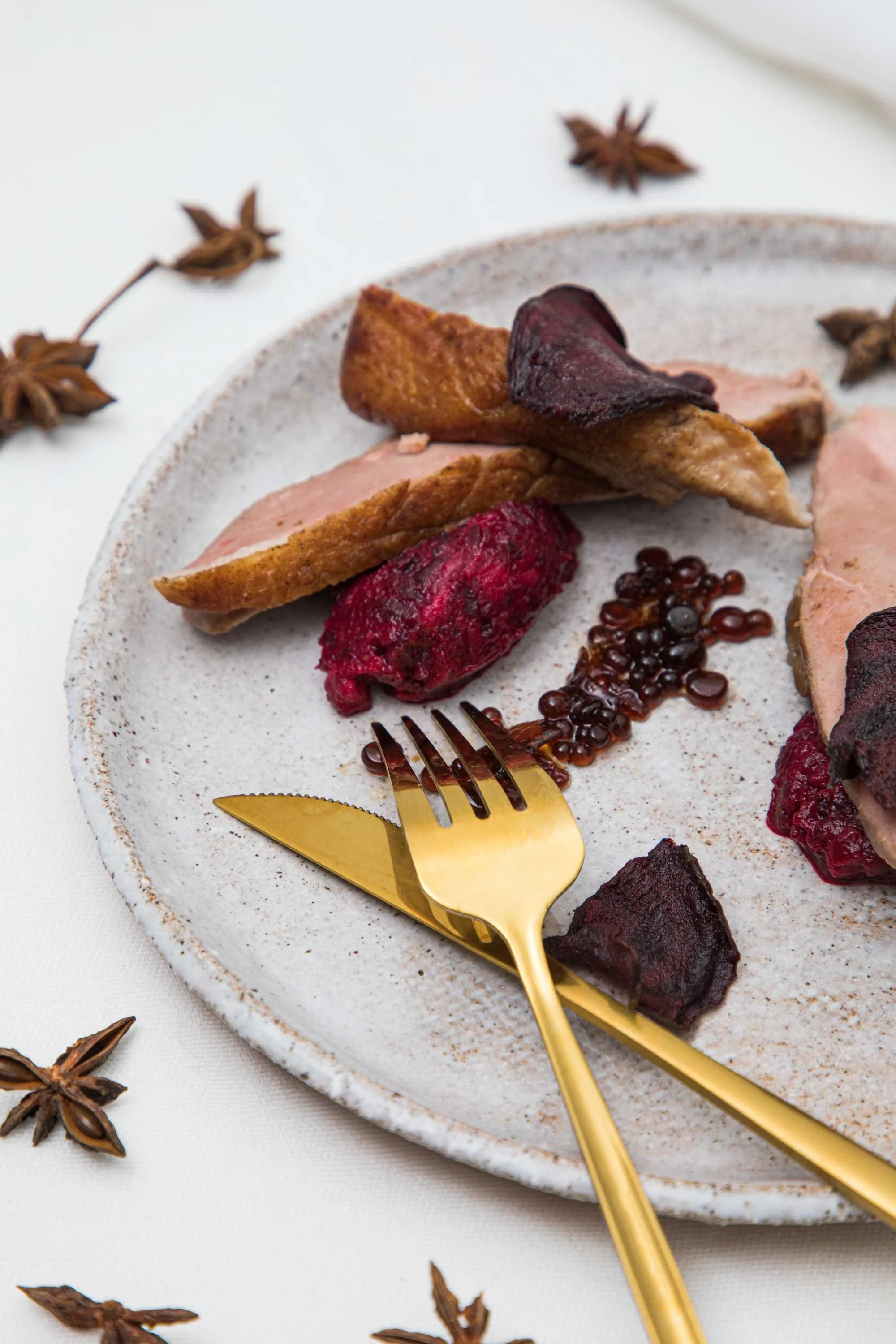 A white ceramic plate with assorted roasted vegetables and berries, accented with a gold fork and knife, on a white tablecloth surrounded by star anise spices.
