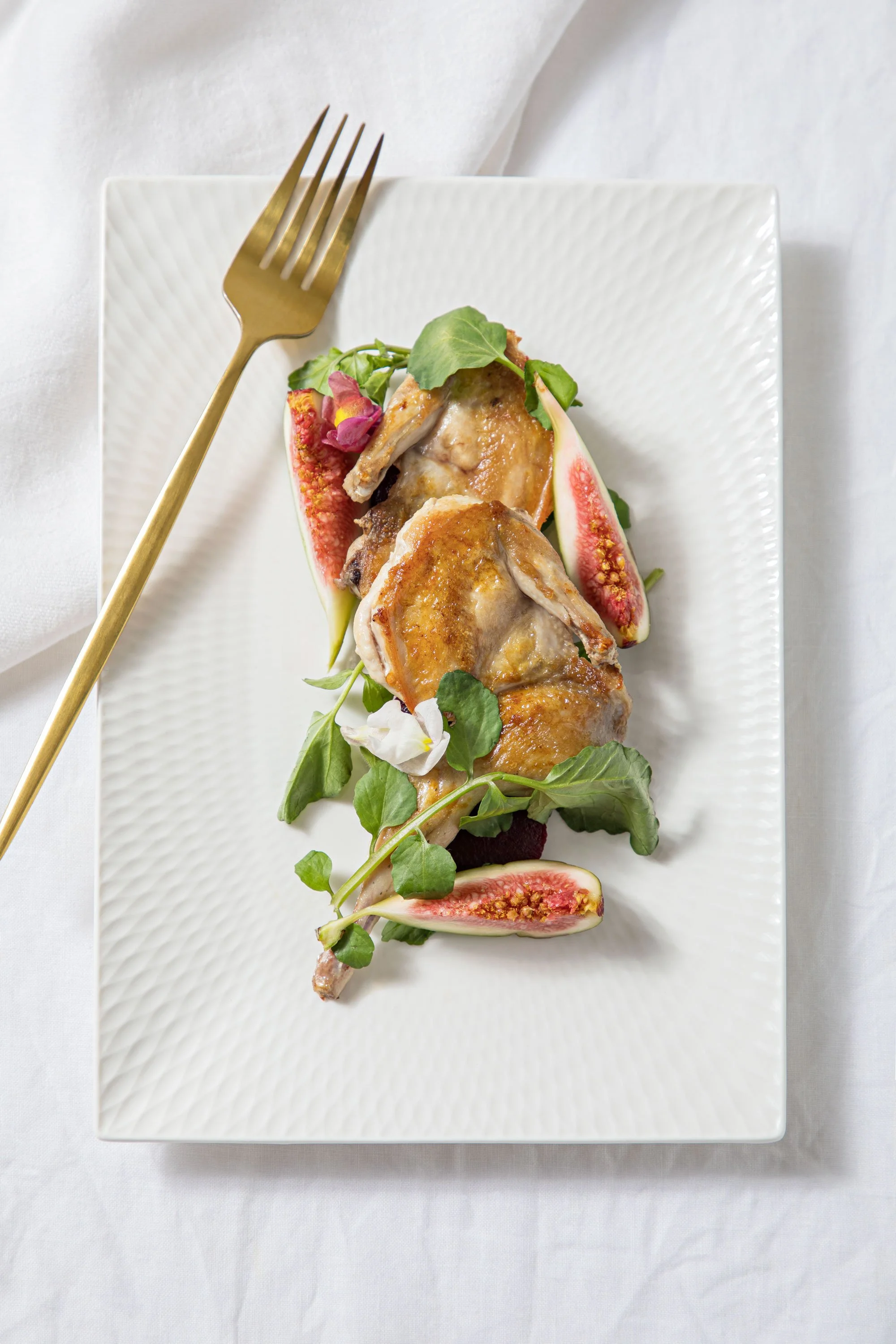 A rectangular white plate with roasted chicken pieces, fig halves, fresh green leaves, and edible flowers, arranged for serving.
