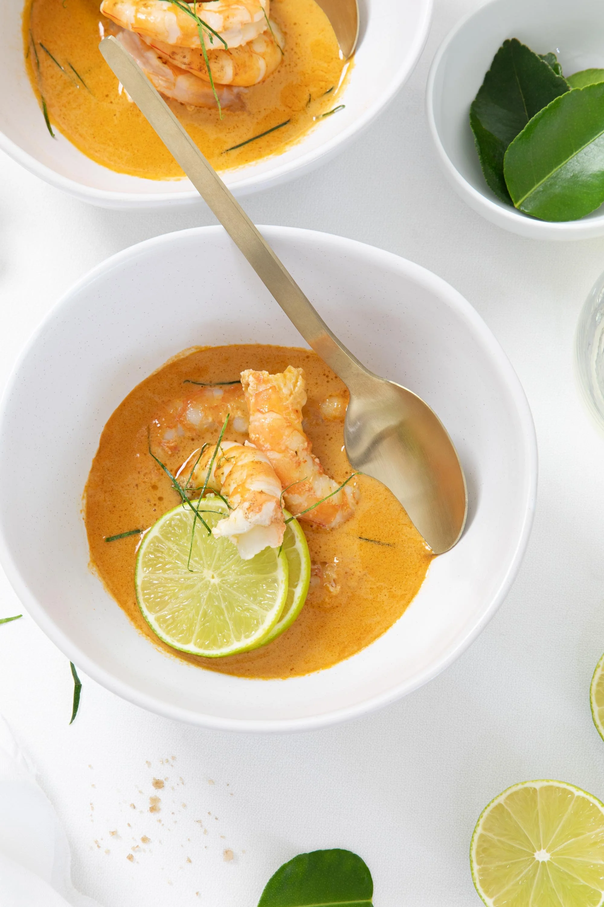 Bowls of shrimp in a creamy orange sauce garnished with lemon slices and herbs, with lime wedges and green leaves on a white surface.