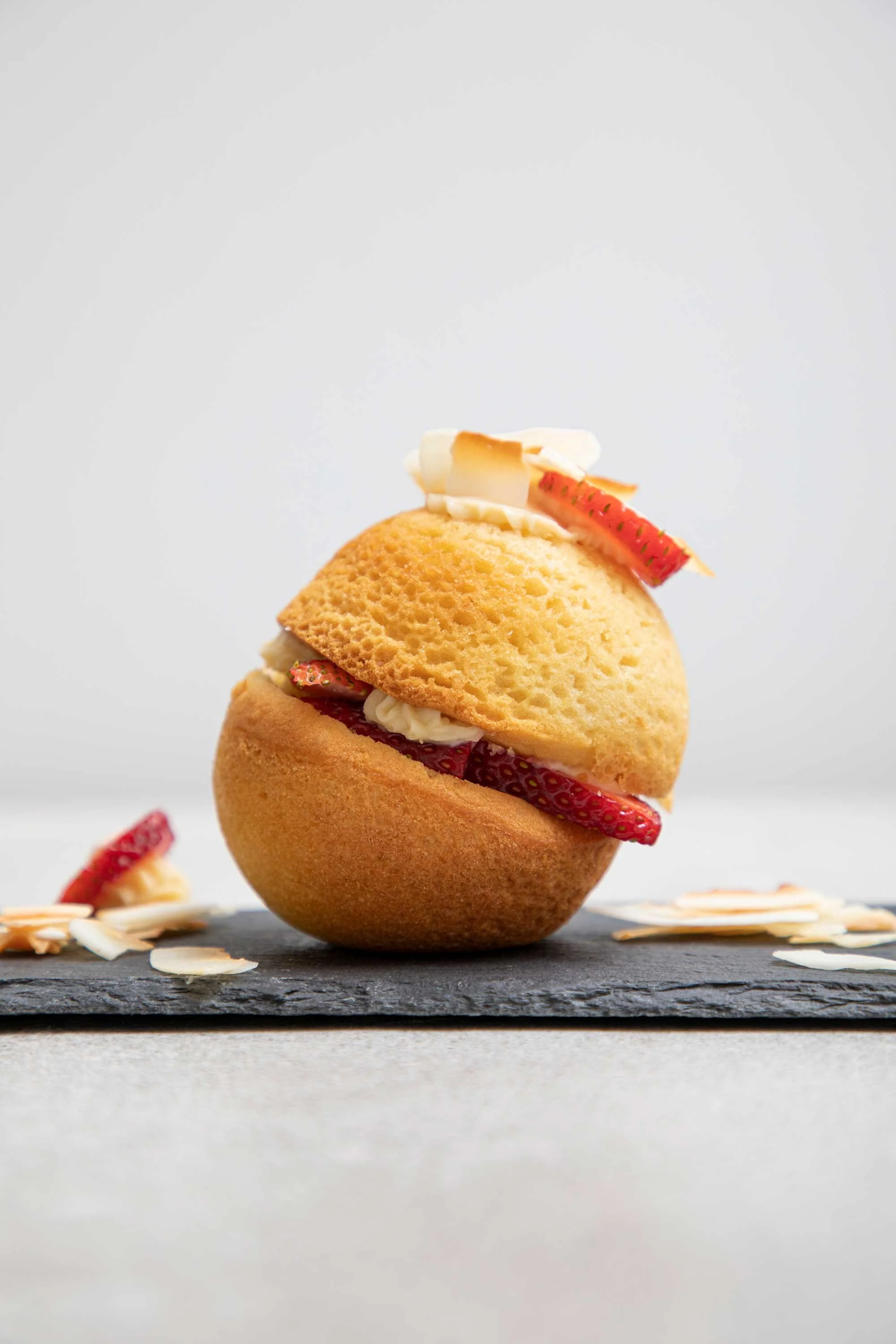 A dome-shaped sponge cake filled with strawberries and cream, topped with sliced strawberries and white chocolate shavings, on a dark slate serving board.
