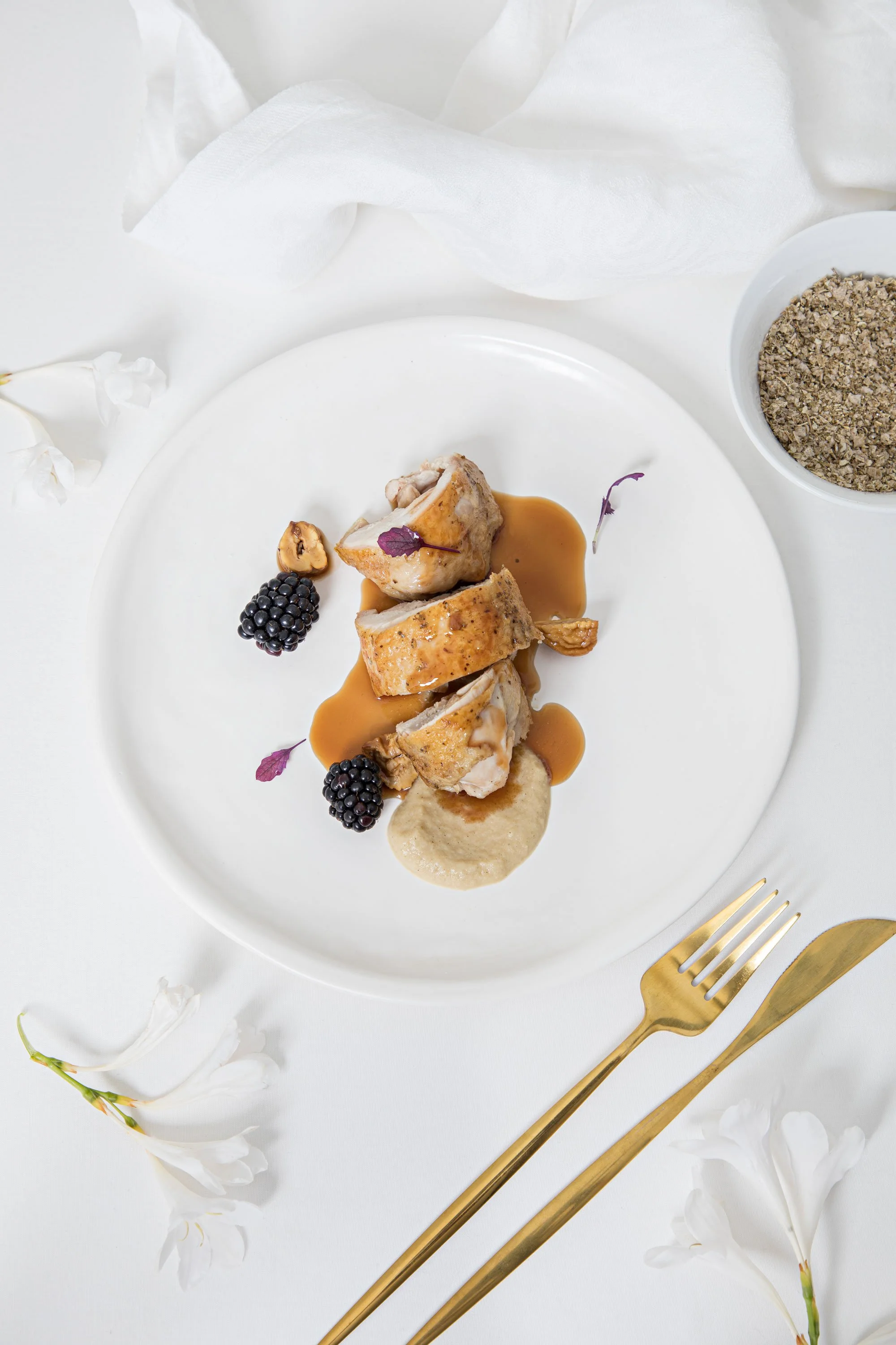 Plate of roasted chicken garnished with purple leaves, blackberries, and sauces, on white table with gold utensils and white flowers.