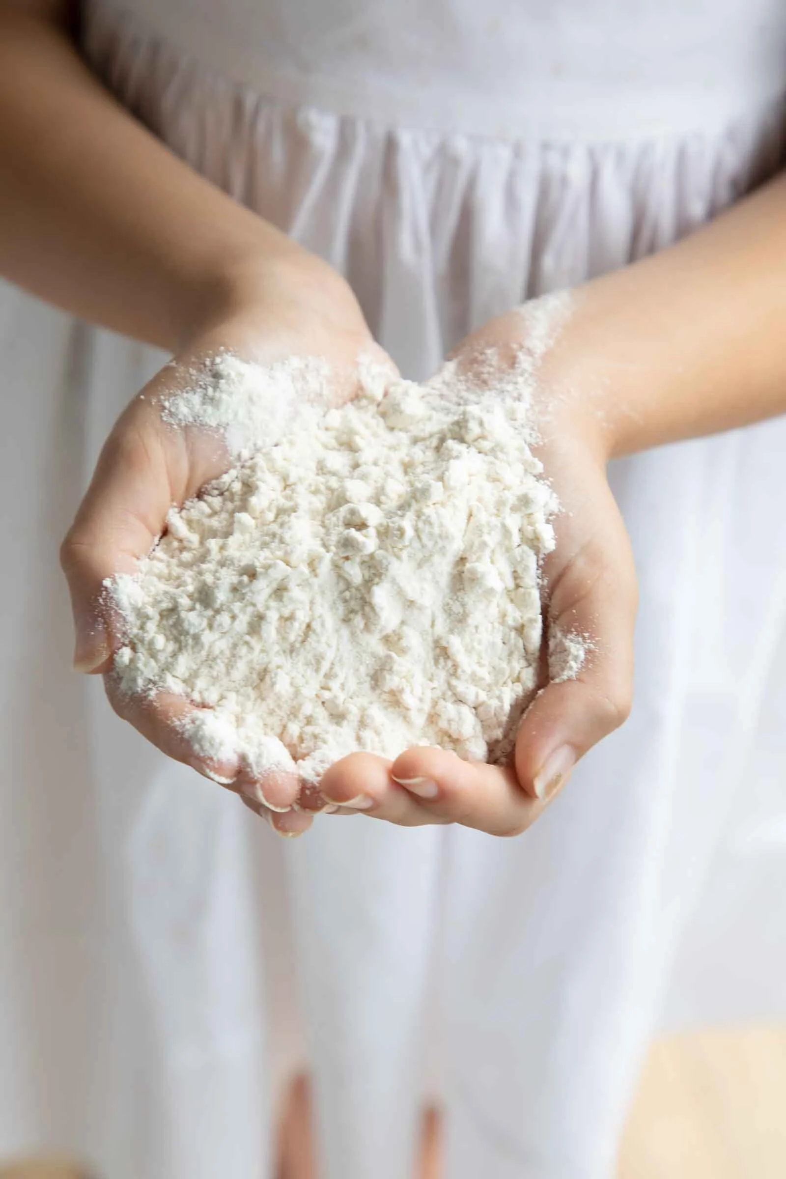 Person holding a handful of white flour, seen from the waist up, dressed in a white apron.