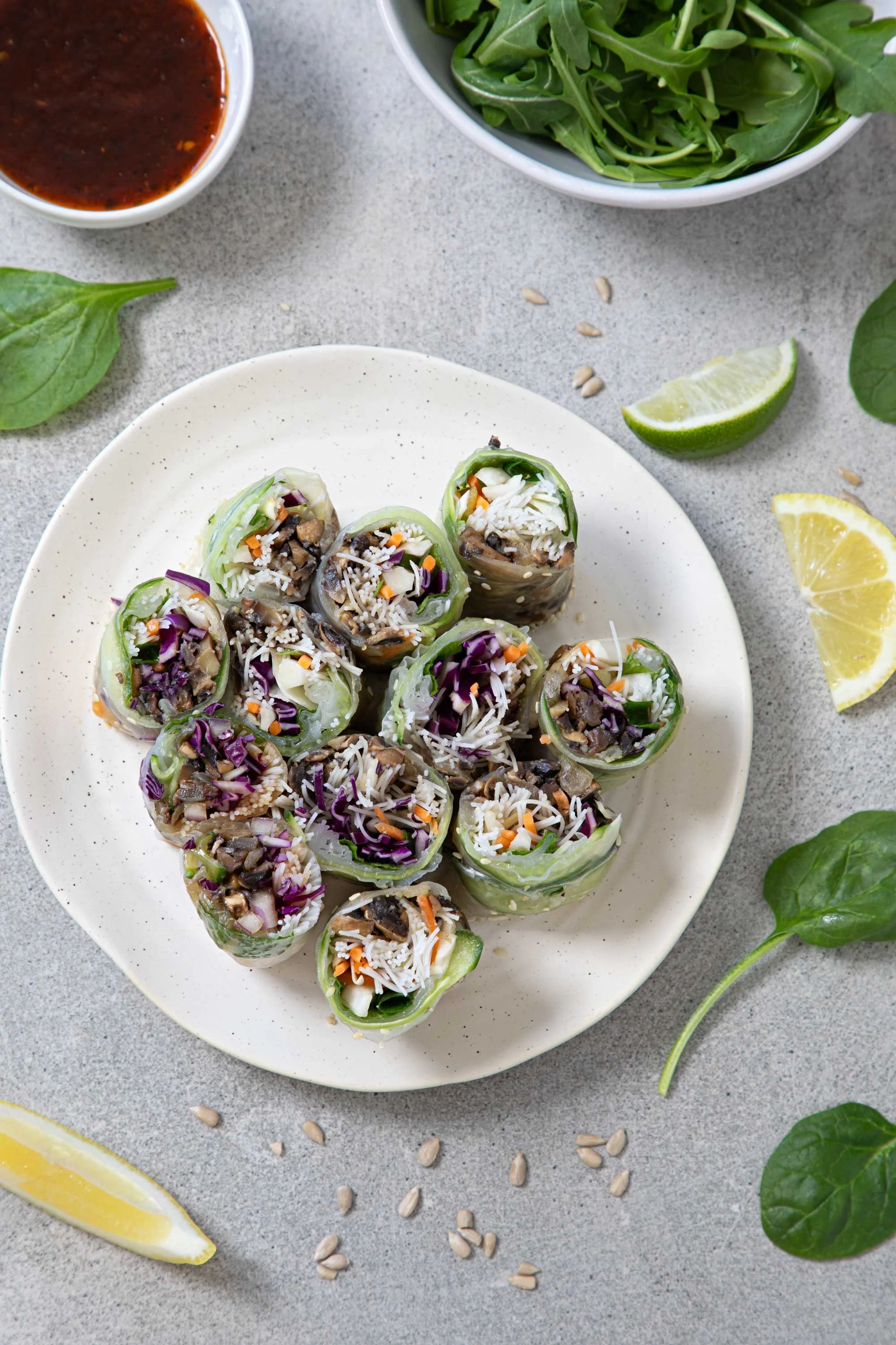 Plate of vegetable spring rolls with shredded vegetables, herbs, and rice noodles, garnished with sunflower seeds and accompanied by lime wedges, on a gray surface with scattered sunflower seeds, fresh spinach, and a small bowl of dipping sauce.