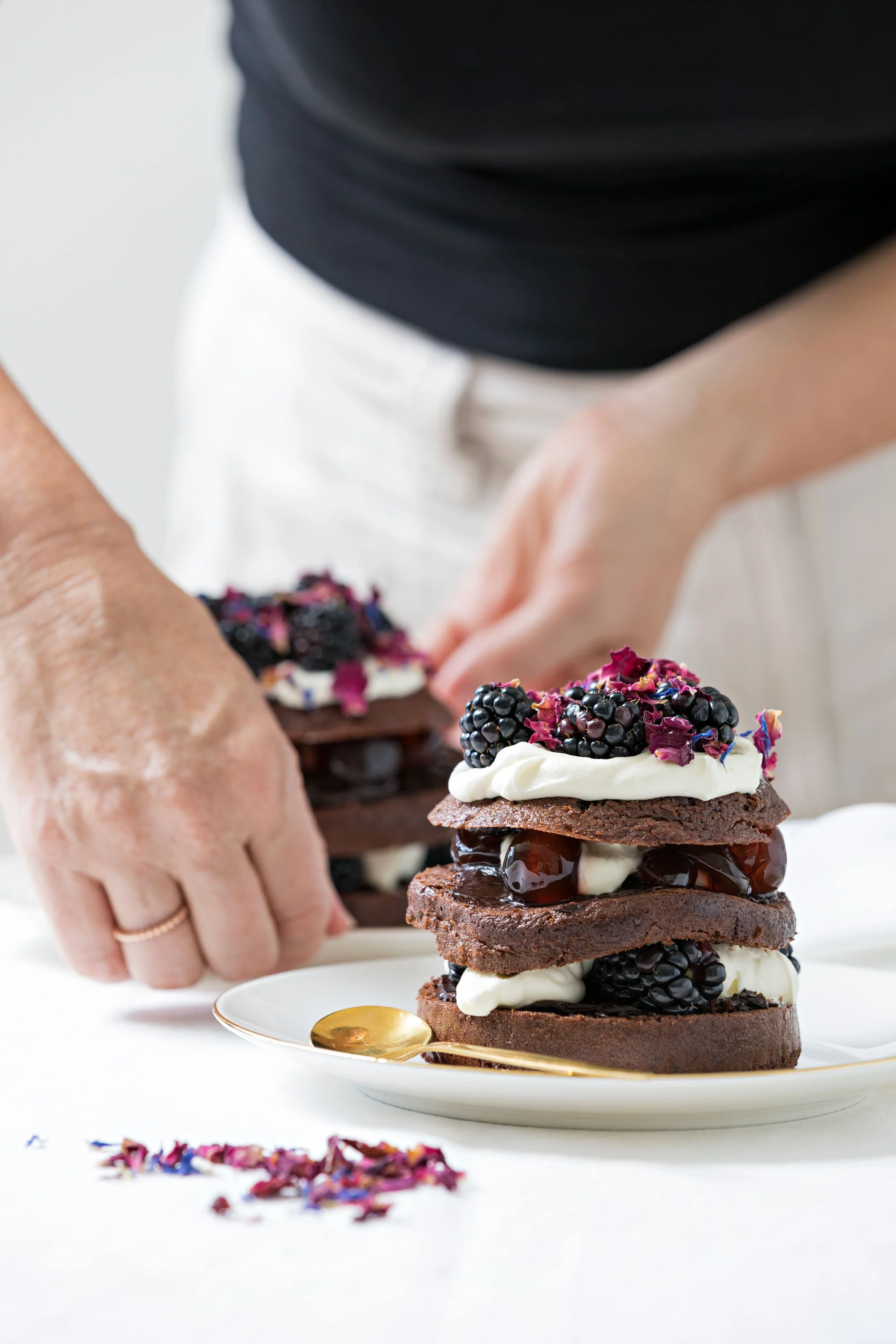 A person assembling a layered chocolate dessert with blackberries, whipped cream, and edible flowers on top, on a white plate with a gold spoon.