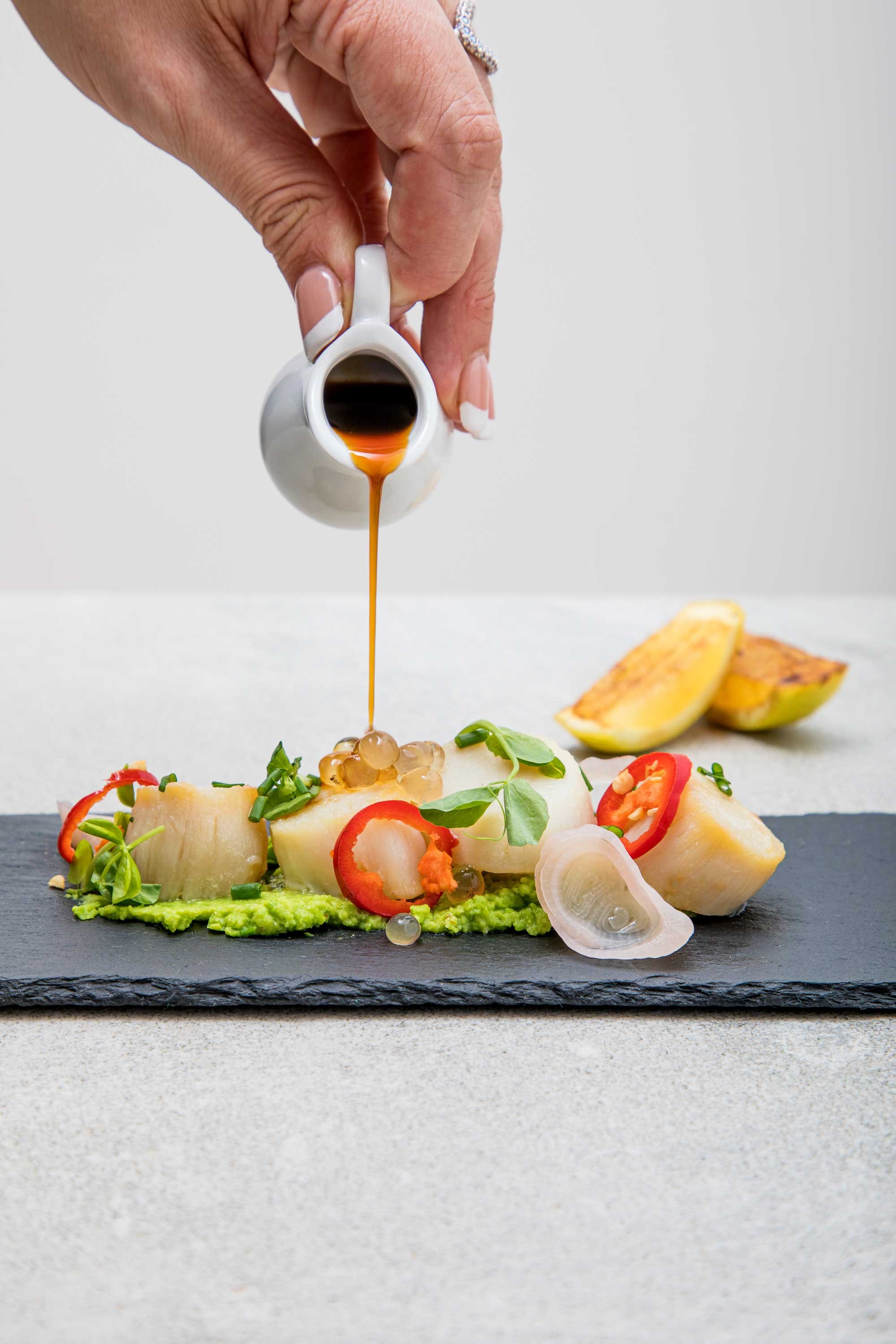 A hand pouring dark liquid sauce over a gourmet dish featuring potatoes, cherry tomatoes, onions, microgreens, and a bright green puree on a black slate plate.