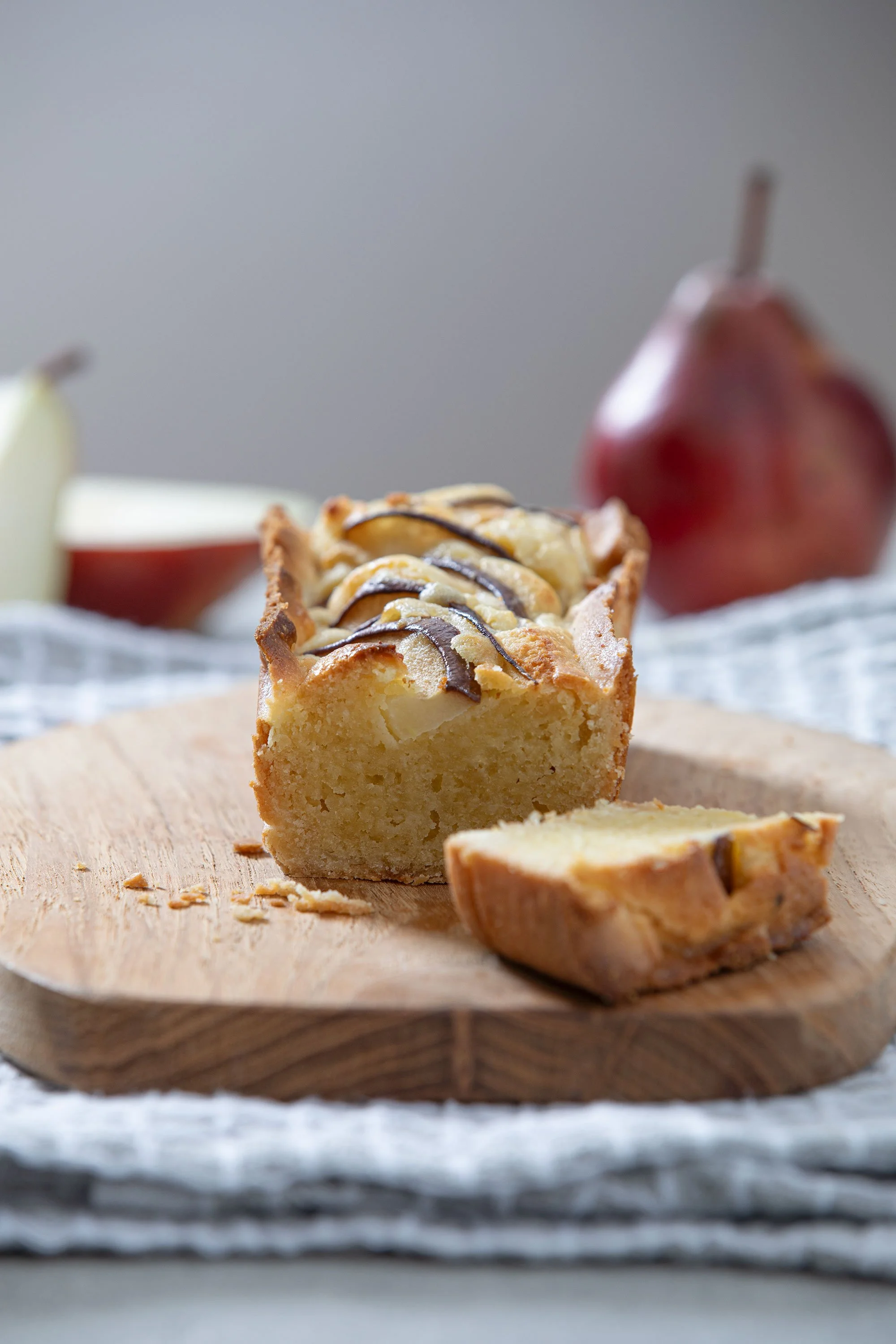 A slice of apple dessert cake with a golden crust, topped with sliced apples and drizzled with chocolate, sitting on a round wooden cutting board. There are whole apples in the background.